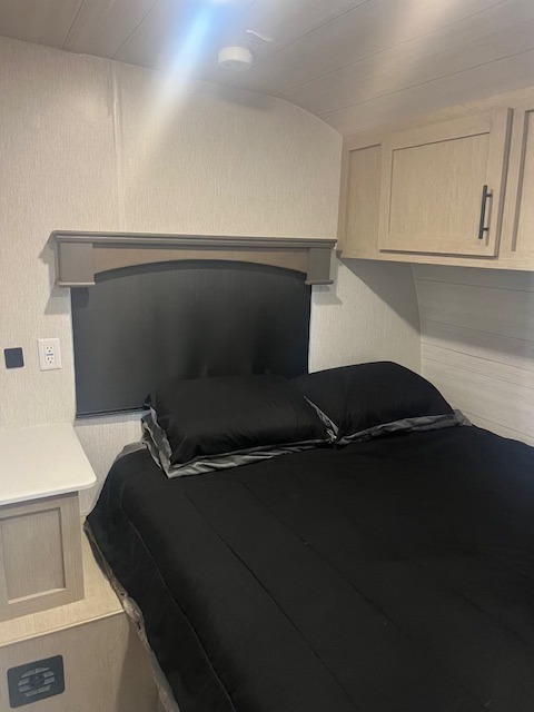 This RV rental bedroom features a comfy bed with black bedding, bedside table, and overhead storage, creating a cozy interior space.. Forest River Shockwave 2023