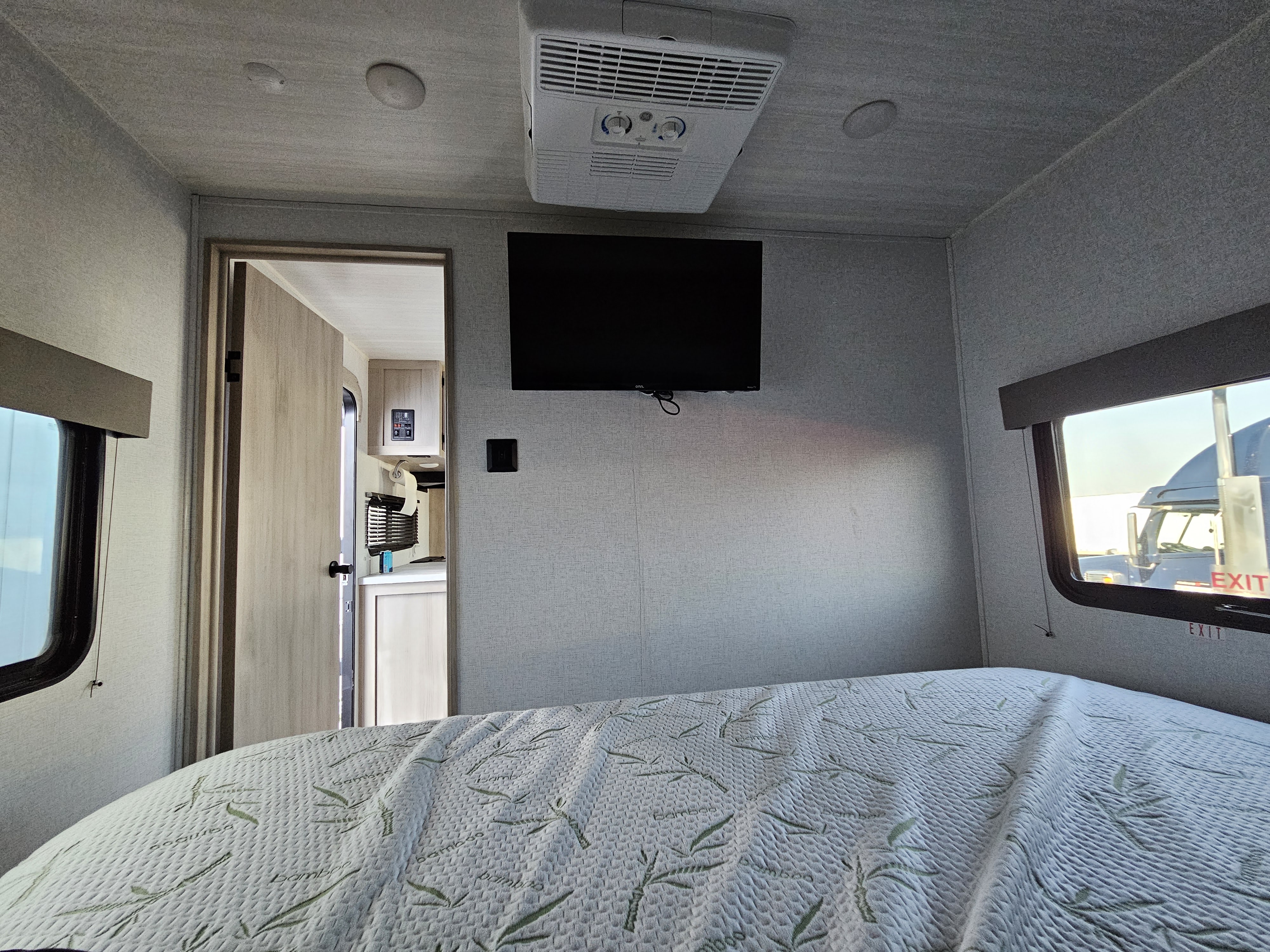 This RV rental bedroom features a patterned bed, wall-mounted TV, ceiling AC unit, and a doorway leading to the main living area.. East to West 271bh 2024