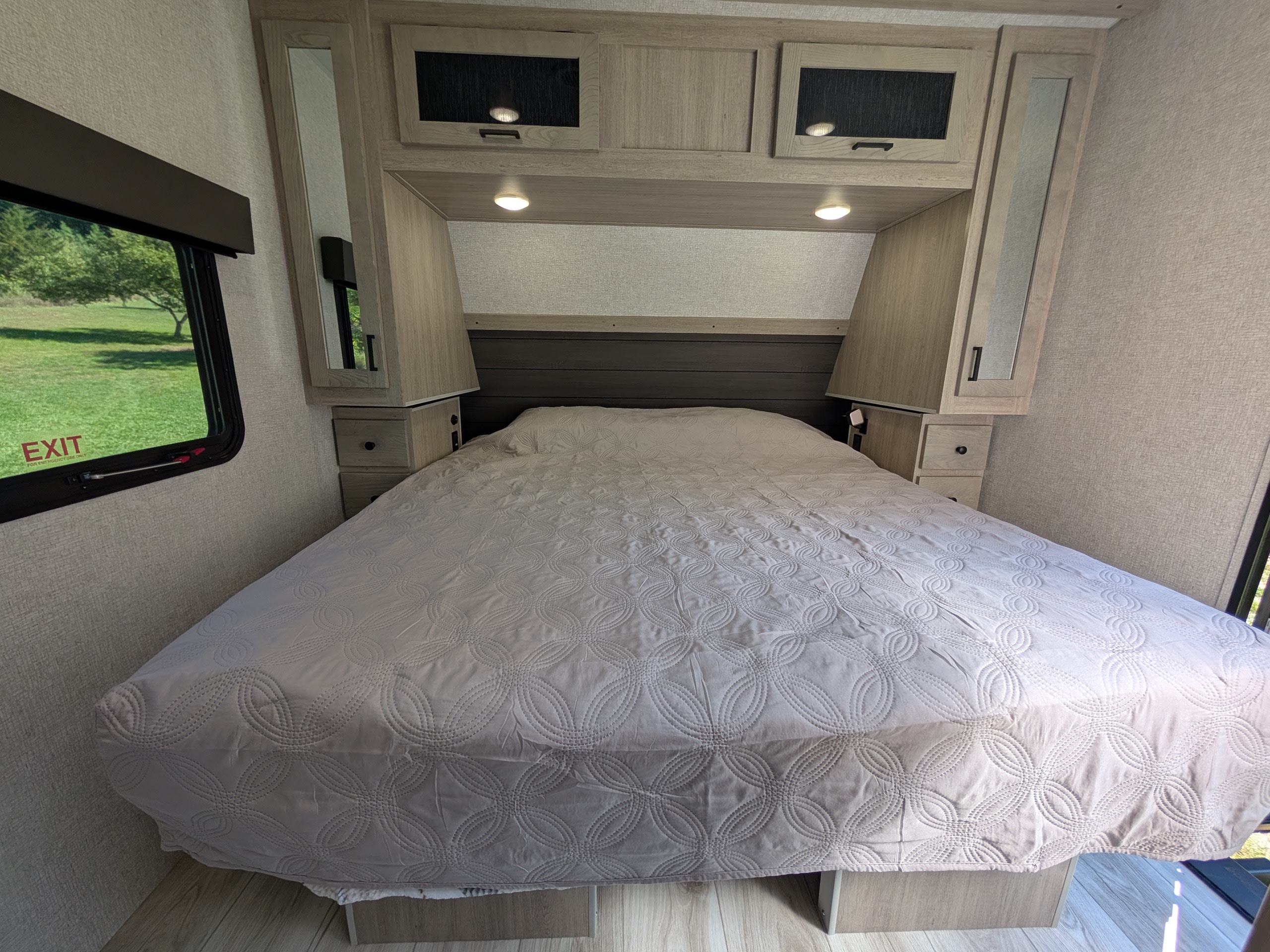 This RV rental bedroom offers a comfortable bed with a quilted cover, overhead cabinets, and a window with a green outdoor view.. Forest River Alta 2850KRL 2025