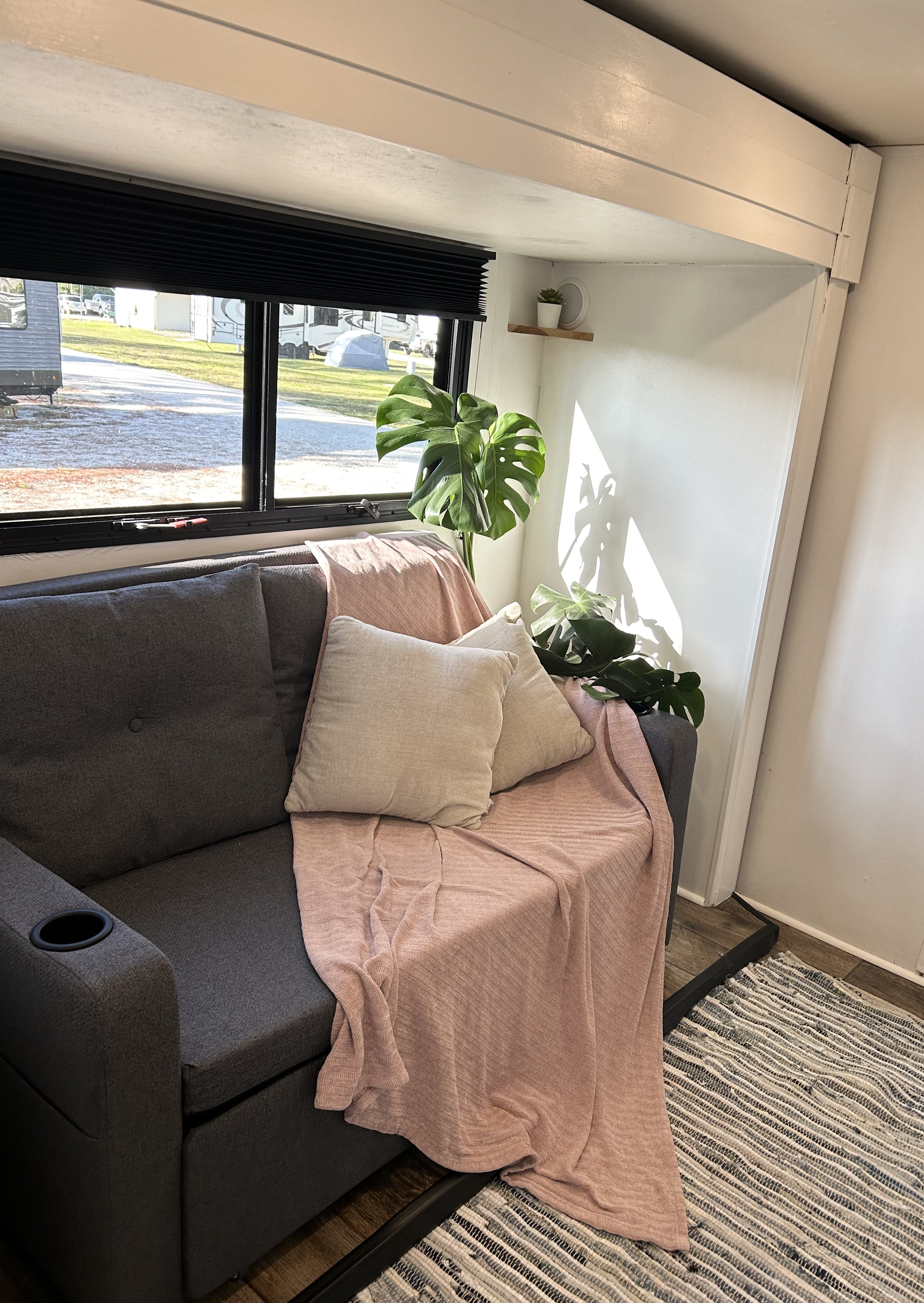 Cozy RV rental interior: A gray couch with pink blanket, Monstera plant, and window view of an RV park.. Highland Ridge RV Roamer 2017