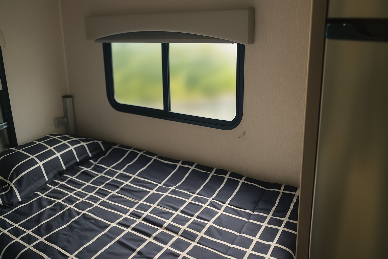 This RV rental's cozy bedroom features a bed with dark blue grid bedding under a window showing a blurry green view.. Coachman Freelander 22 XG 2023