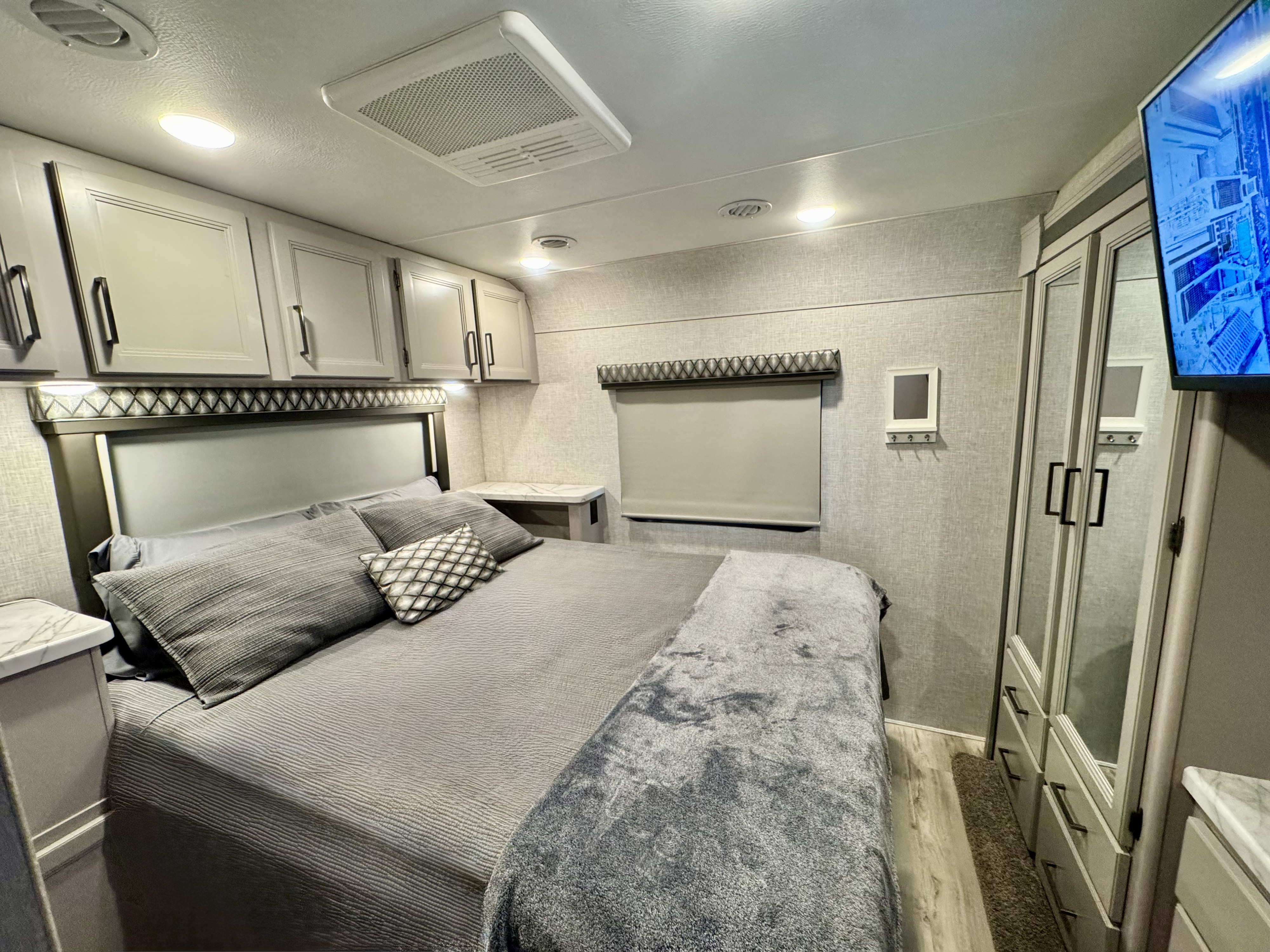 Step inside this cozy, modern RV rental bedroom featuring a large gray bed, overhead cabinets, and a mounted TV.. Thor Geneva 2023