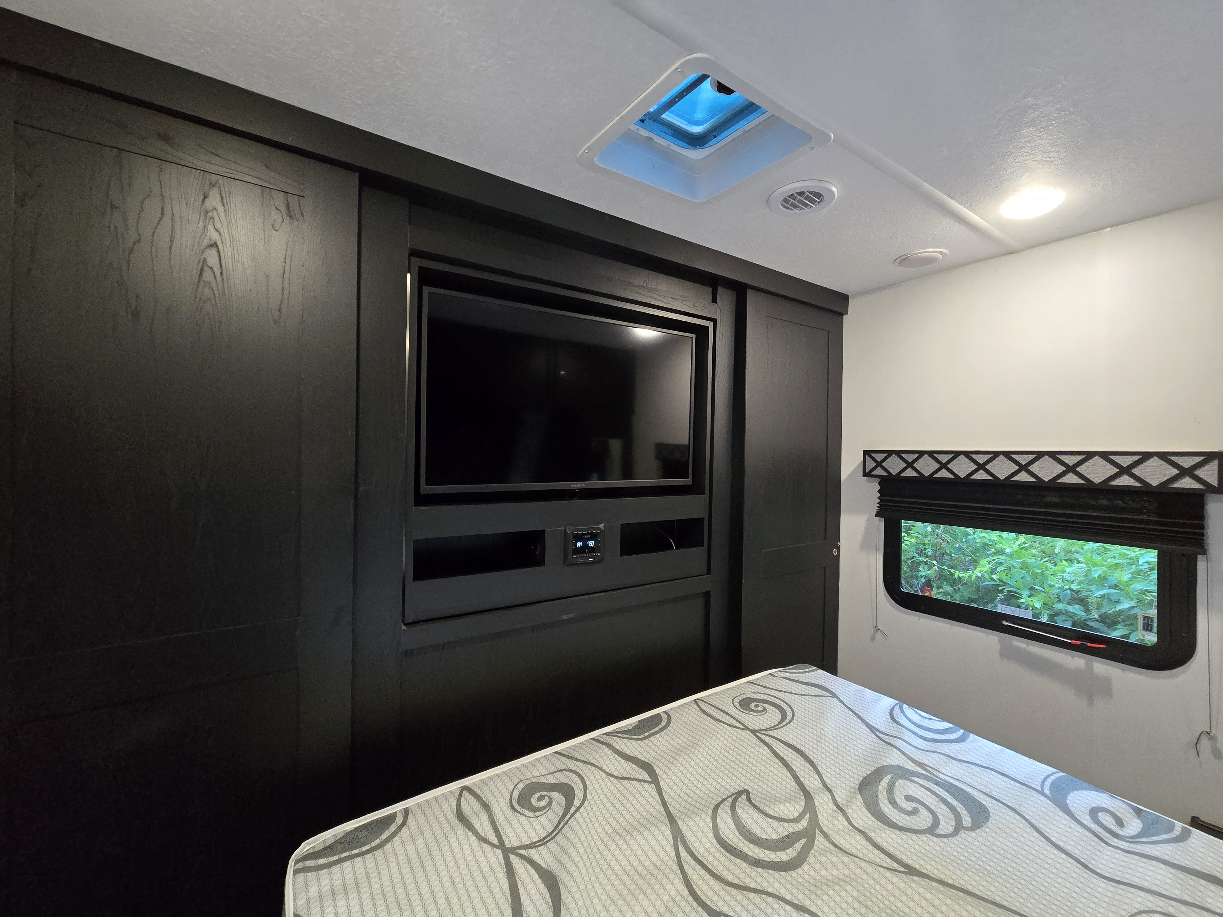RV rental bedroom: dark wood entertainment unit with a TV and stereo, patterned bed, and window looking out to greenery.. Forest River Coachmen Freedom Express Ultra Lite 2022