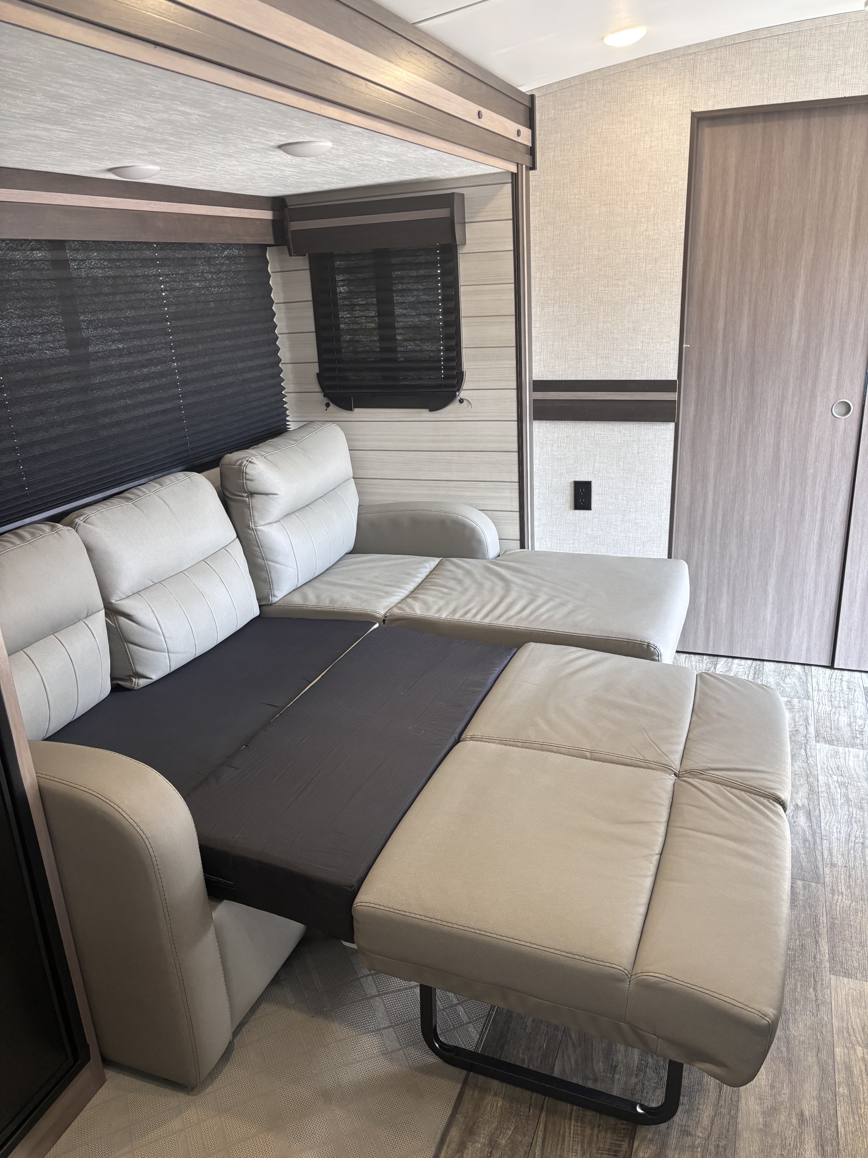 An rv rental's interior with a beige convertible sofa, partially extended into a bed, alongside windows with blinds and a door.. Crossroads RV Sunset Trail Super Lite 2022