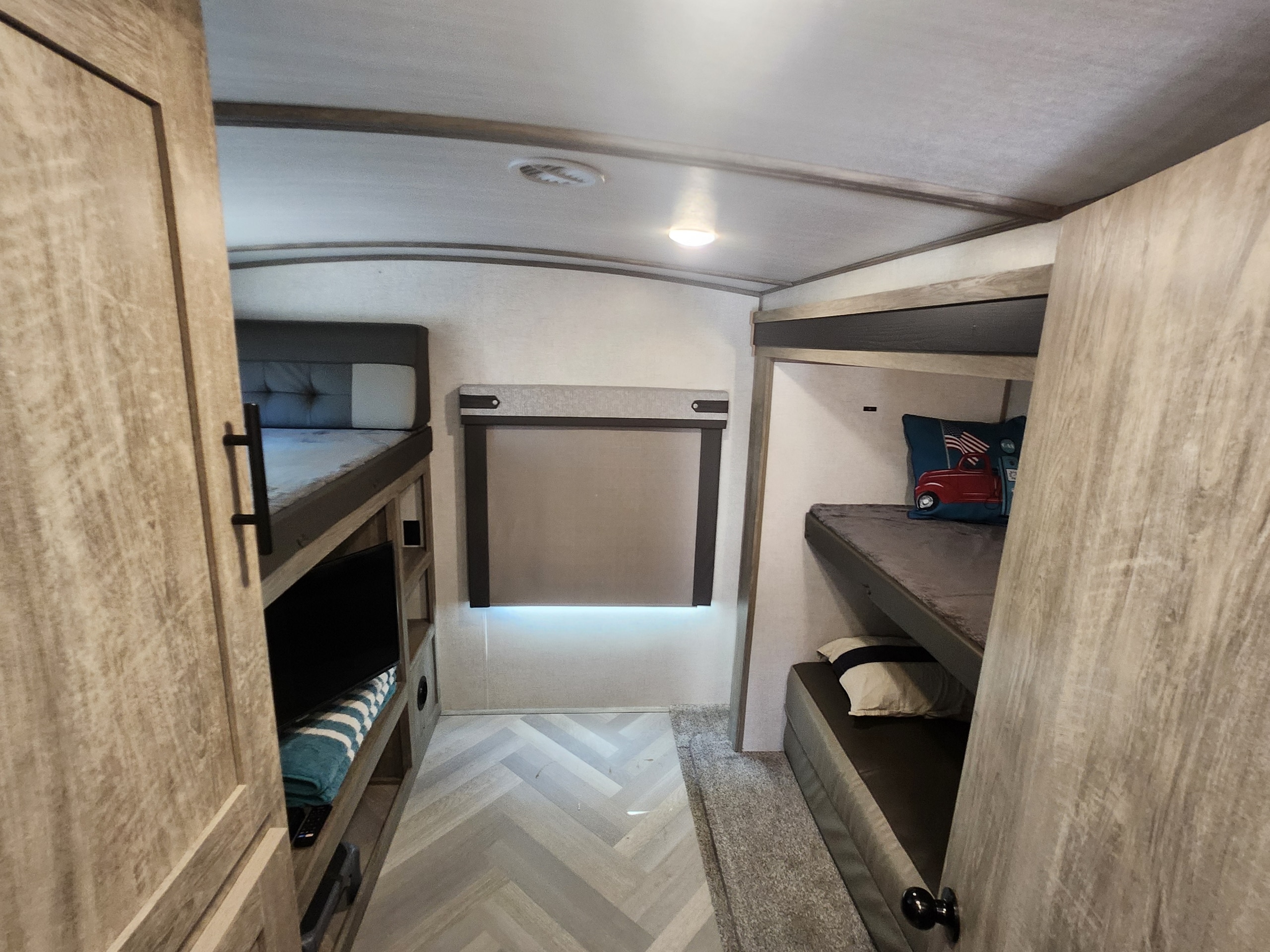 Cozy RV rental bunk room with herringbone flooring and plush carpet. Features left bunks with TV, right bunks with a red truck pillow, and an illuminated window shade.. Forest River Wildwood Heritage Glen 2021