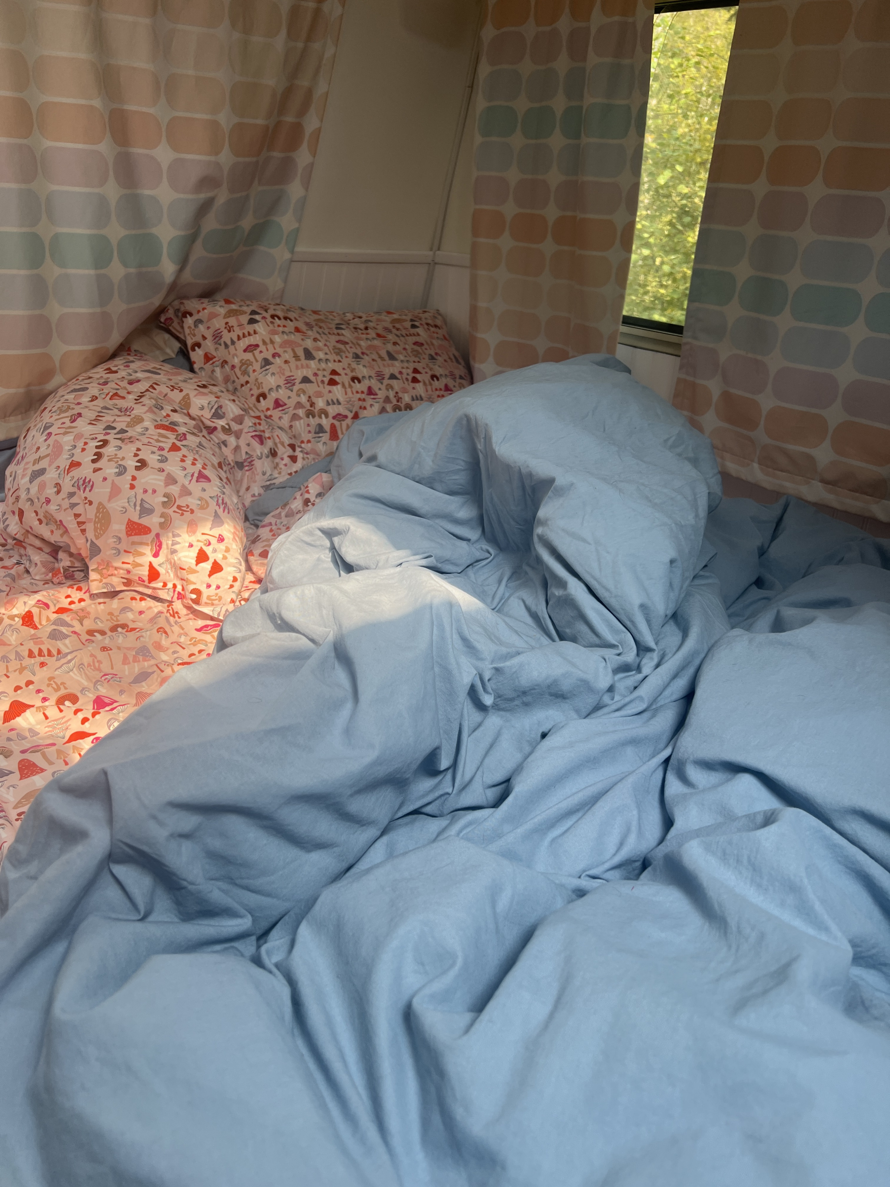 Cozy RV rental interior. An unmade bed with mushroom-patterned pillows and a blue duvet catches morning sunlight near striped curtains.. Ford Econoline 1988