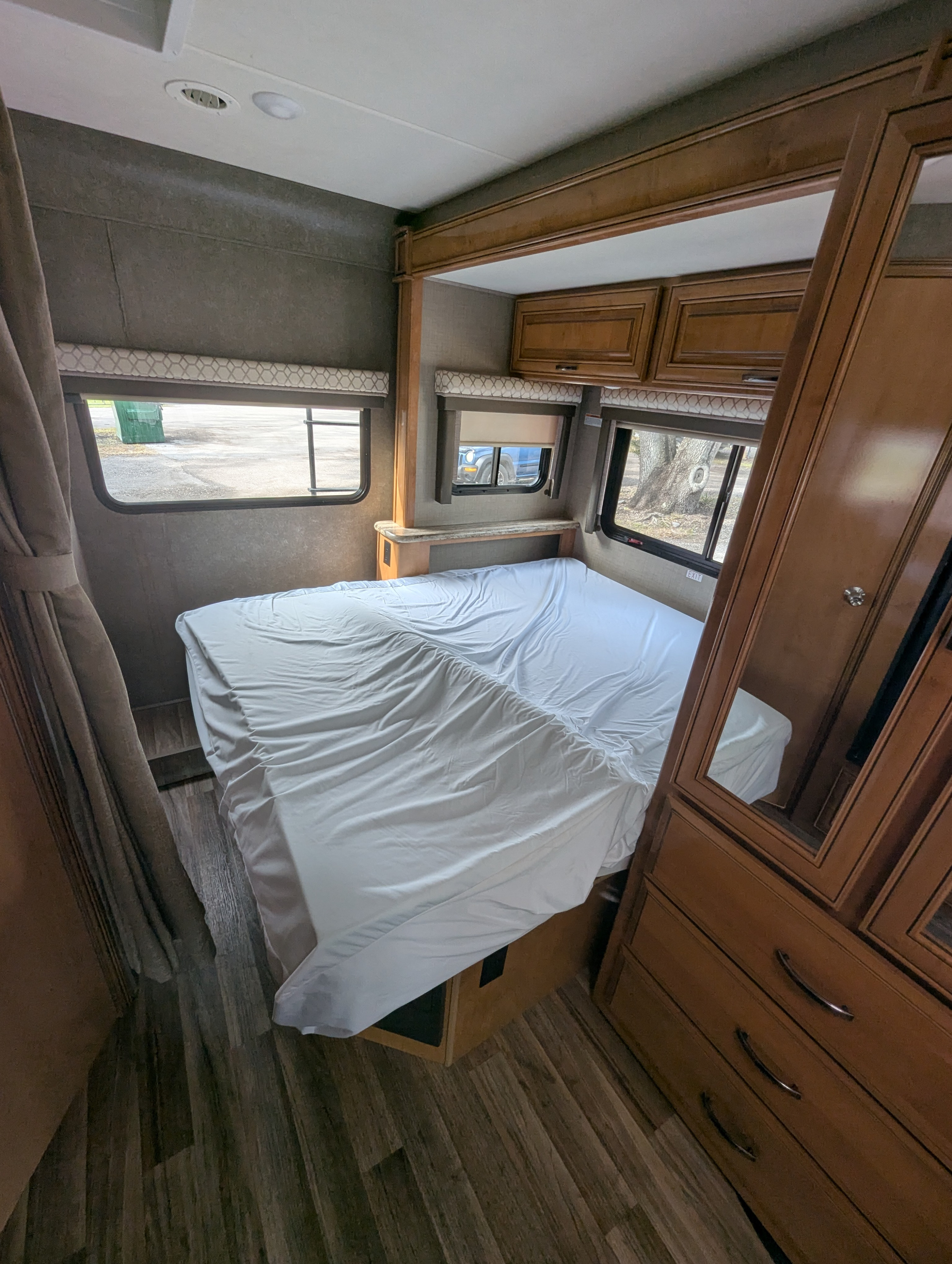 RV rental bedroom featuring a neat bed with white sheets, windows showing outside, wood cabinets & mirrored wardrobe.. Thor Motor Coach Quantum 2018