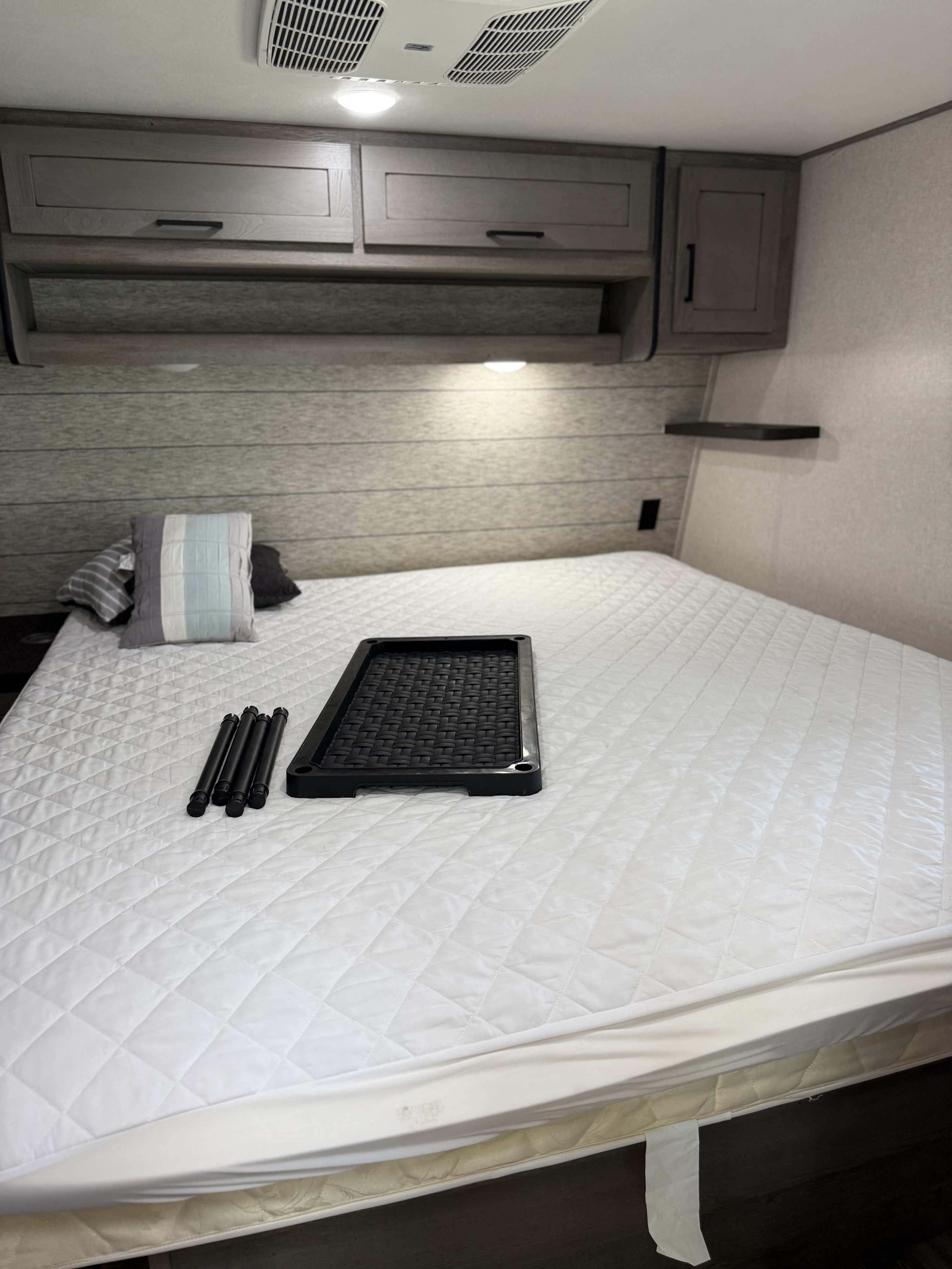 Preparing an RV rental bedroom: quilted mattress, pillows, and unassembled storage tray/legs on the bed. Overhead cabinets & AC.. Keystone RV Raptor Toy Hauler 2021