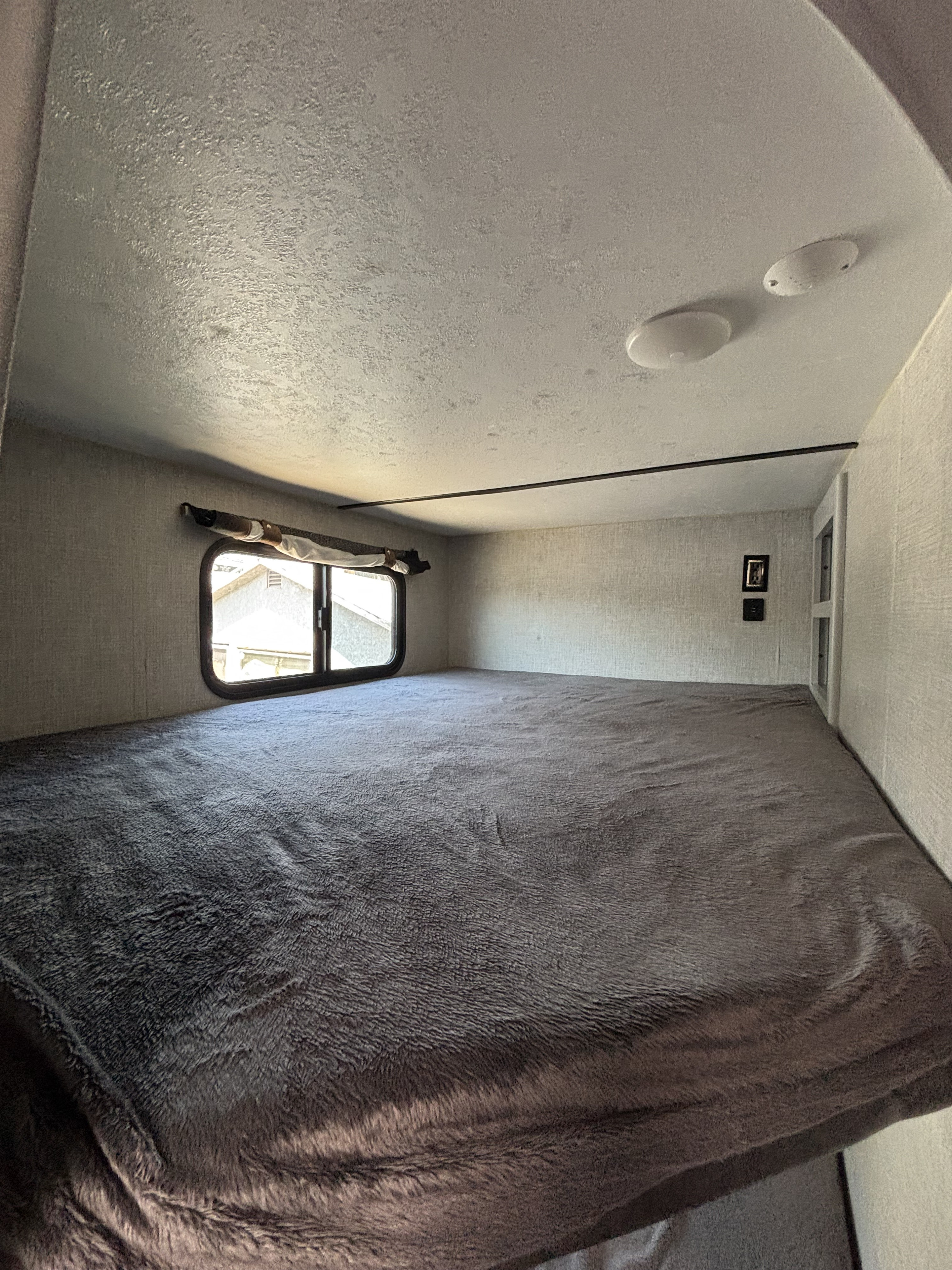 Cozy RV rental bunk bed with a soft, dark grey blanket, window, textured walls, and ceiling lights, ideal for travel.. Keystone RV Bullet Ultra Lite 2021