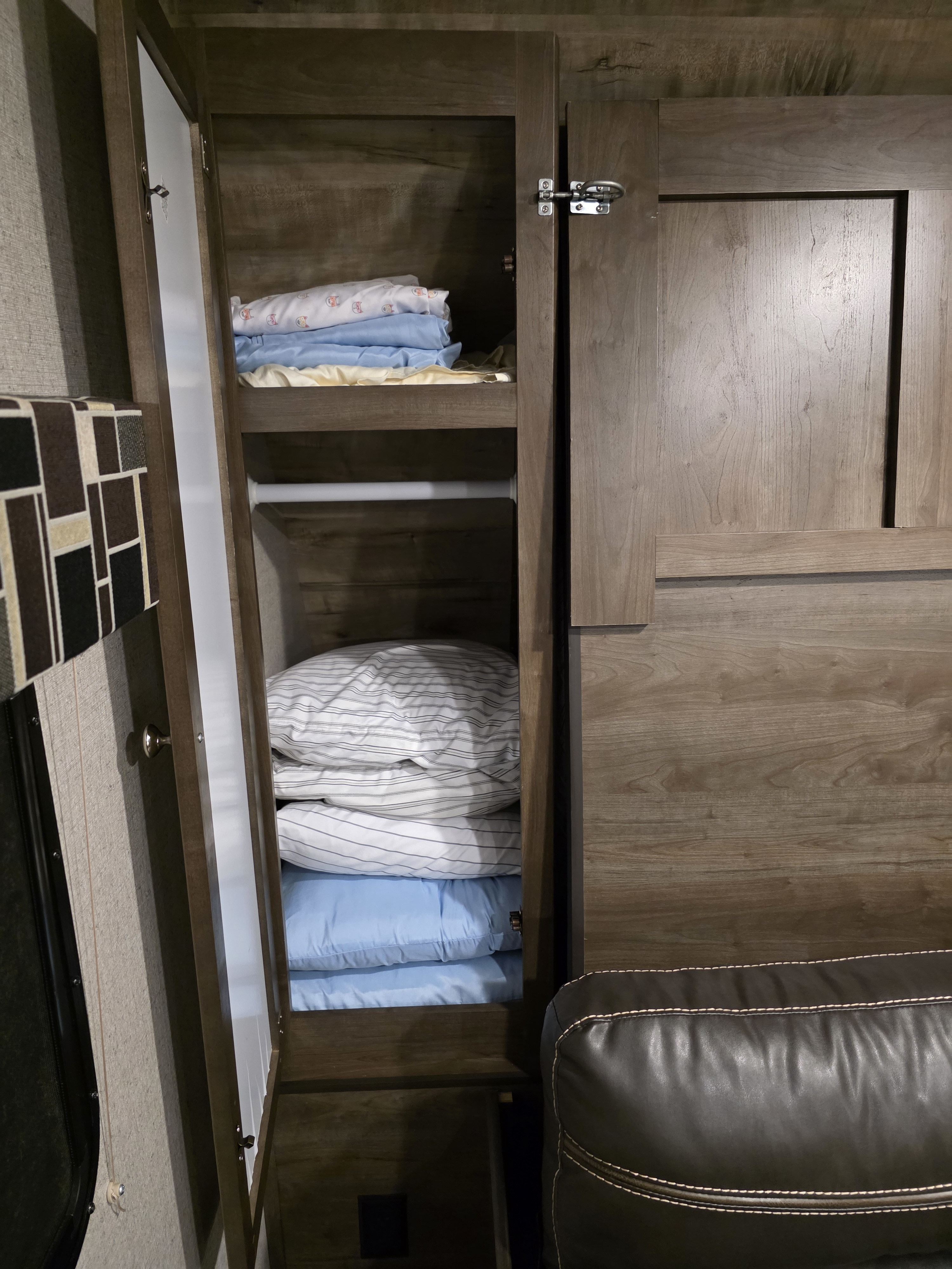 RV rental interior: open wooden closet with stacked bedding & pillows. Efficient storage next to a cozy leather recliner.. Dutchmen Coleman Light LX 2019