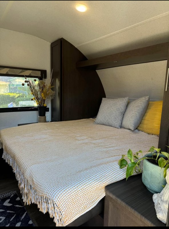 Cozy RV rental bedroom features a striped bedspread, grey & yellow pillows, dark wood cabinetry, and a bright window view.. Forest River Salem Select 2020