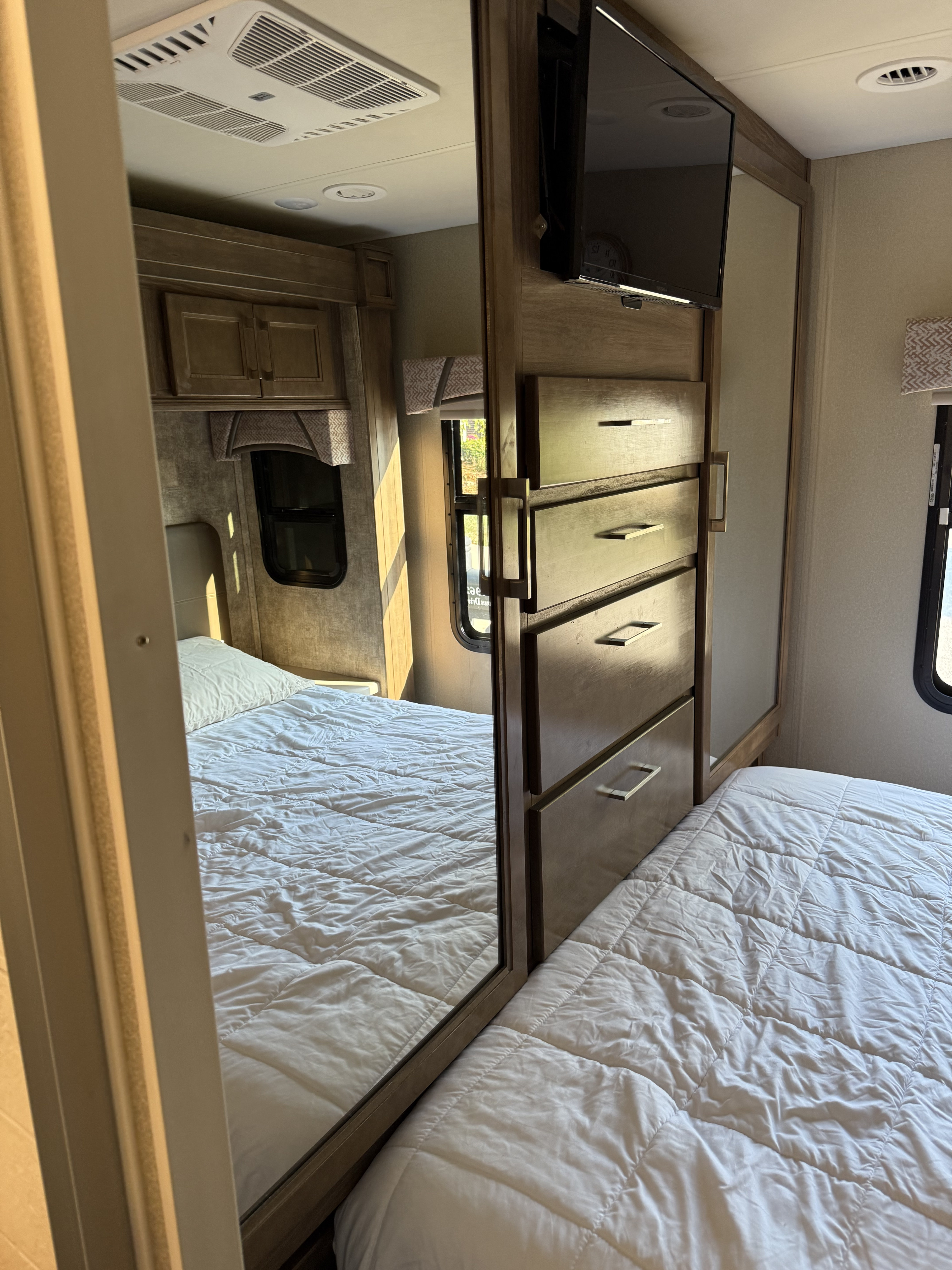 Cozy RV rental bedroom with a comfy white bed, mirrored dresser, and TV. Sunlight streams through the window, highlighting the interior.. Jayco Greyhawk Prestige 2018