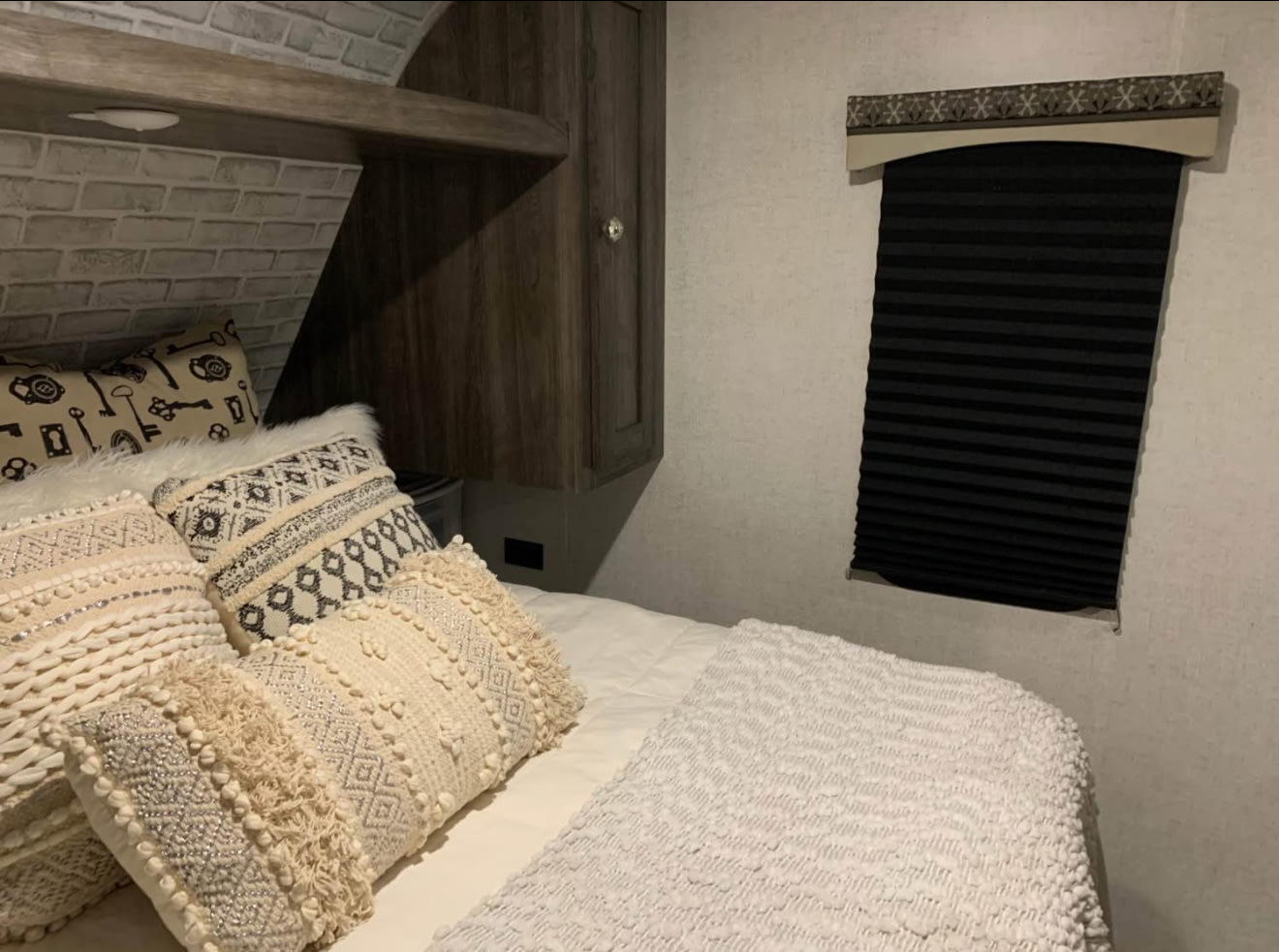 This cozy RV rental bedroom features a bed with plush, patterned pillows, a brick-textured wall, and a window with a dark pleated blind.. Starcraft Autumn Ridge 2020