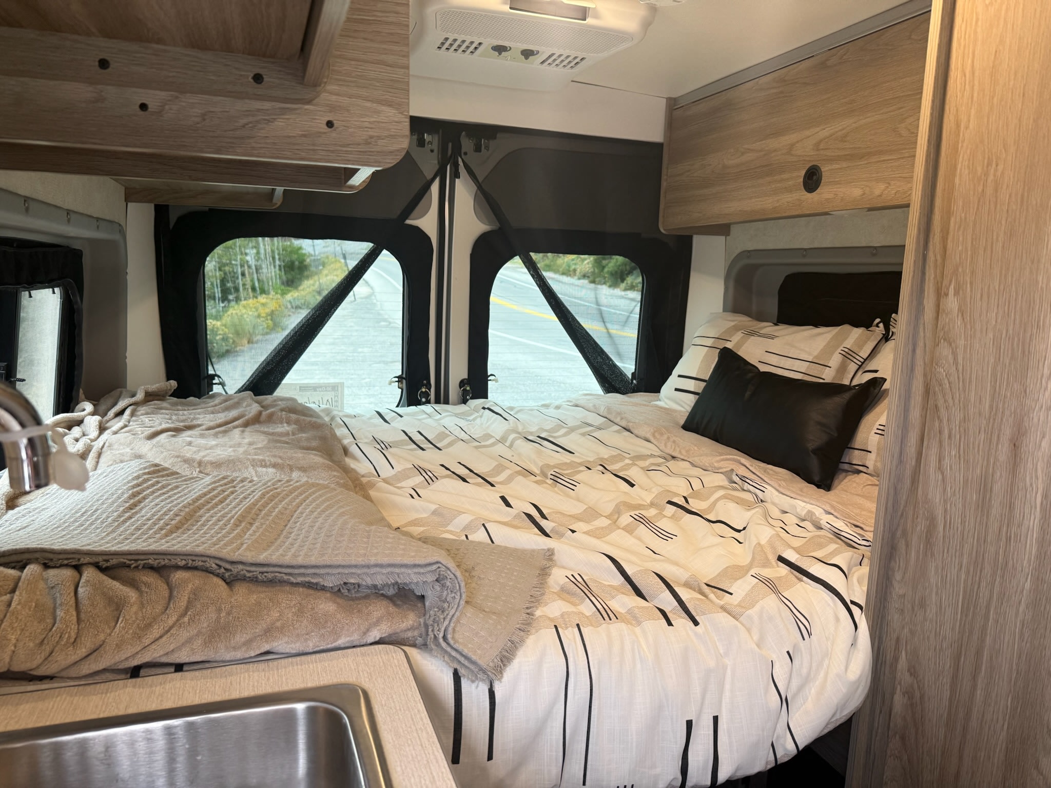 Inside a compact rv rental: cozy bed with graphic bedding, small sink, overhead cabinets, and a scenic view through rear windows.. Winnebago Soilis 2025