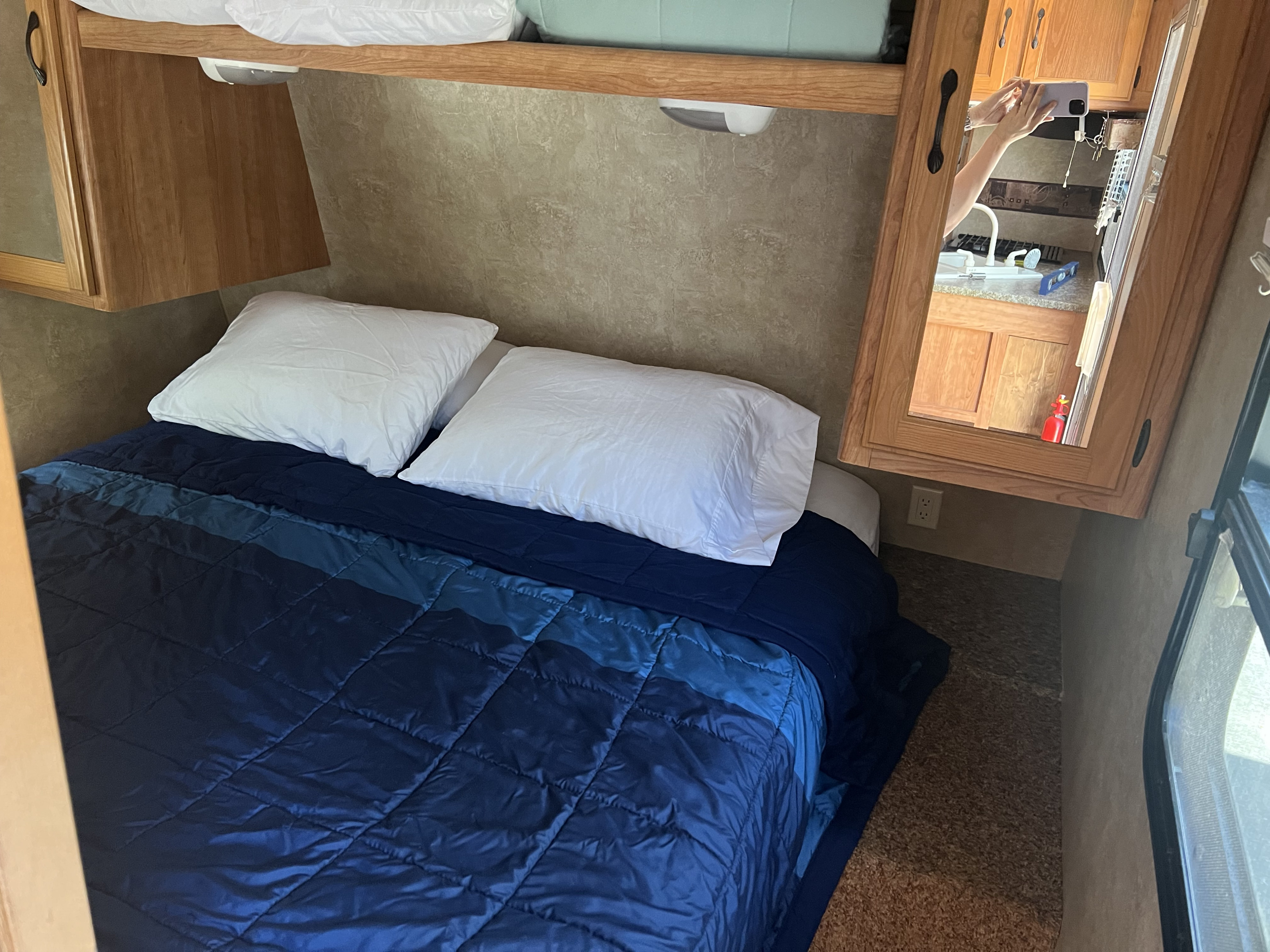 Cozy RV rental bedroom with a comfy bed, blue comforter, and white pillows. Overhead storage and kitchen reflection visible.. Keystone RV Sprinter Select 2010