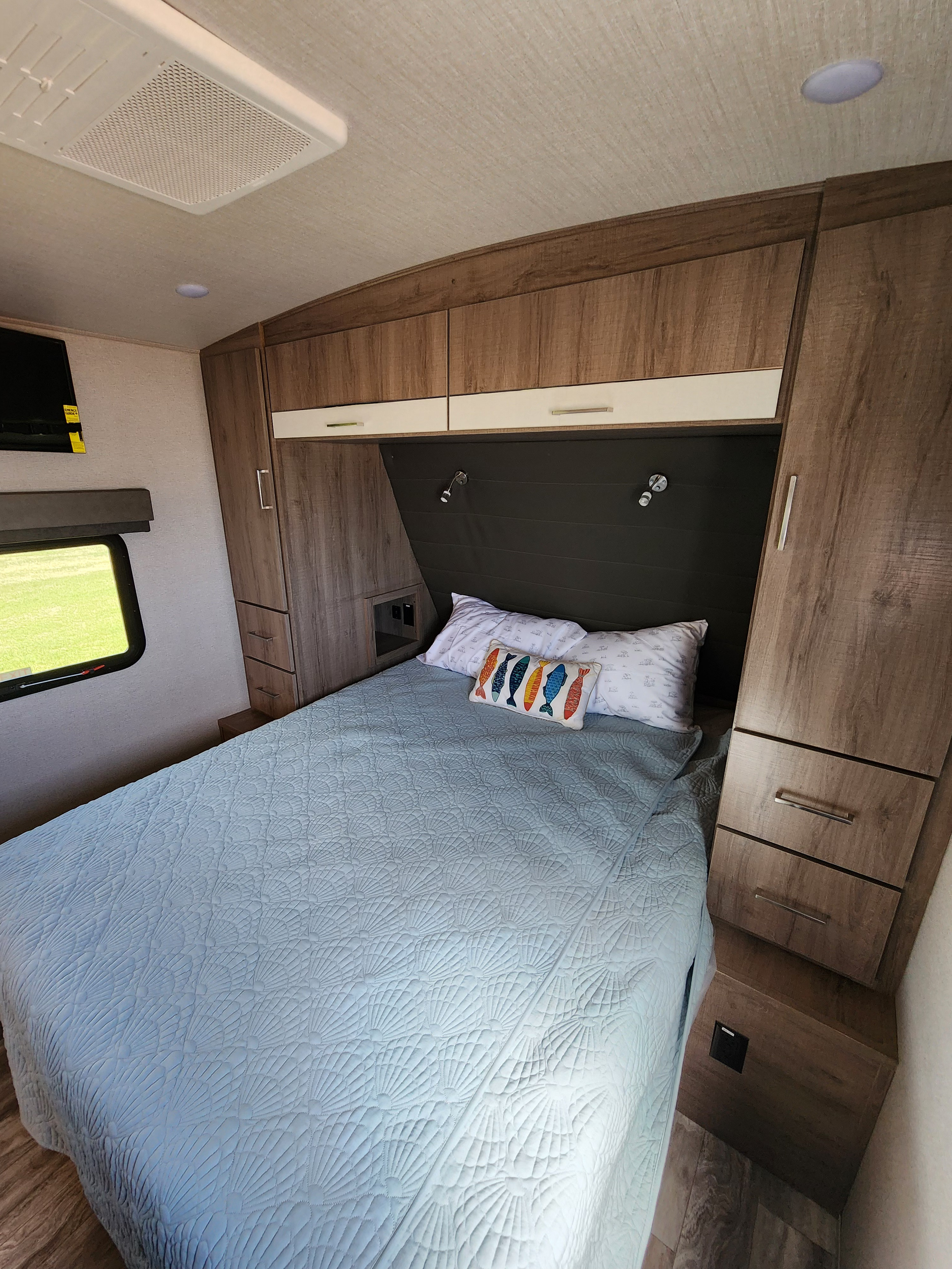 Cozy RV rental bedroom featuring a light blue quilted bedspread, overhead storage, and a bright window with a green view.. Grand Design Imagine 2022