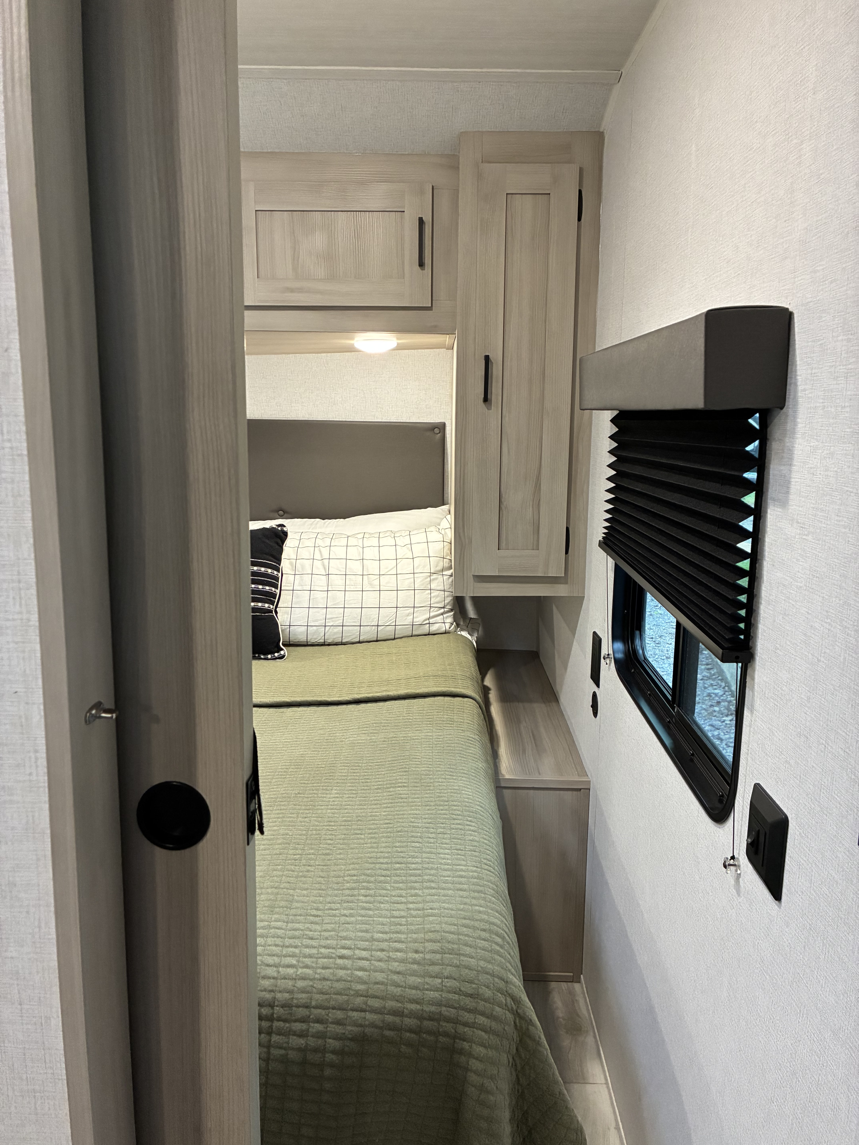 A cozy rv rental bedroom with a bed, green quilt, overhead cabinets, and a window featuring a dark pleated blind.. East To West Della Terra 2025