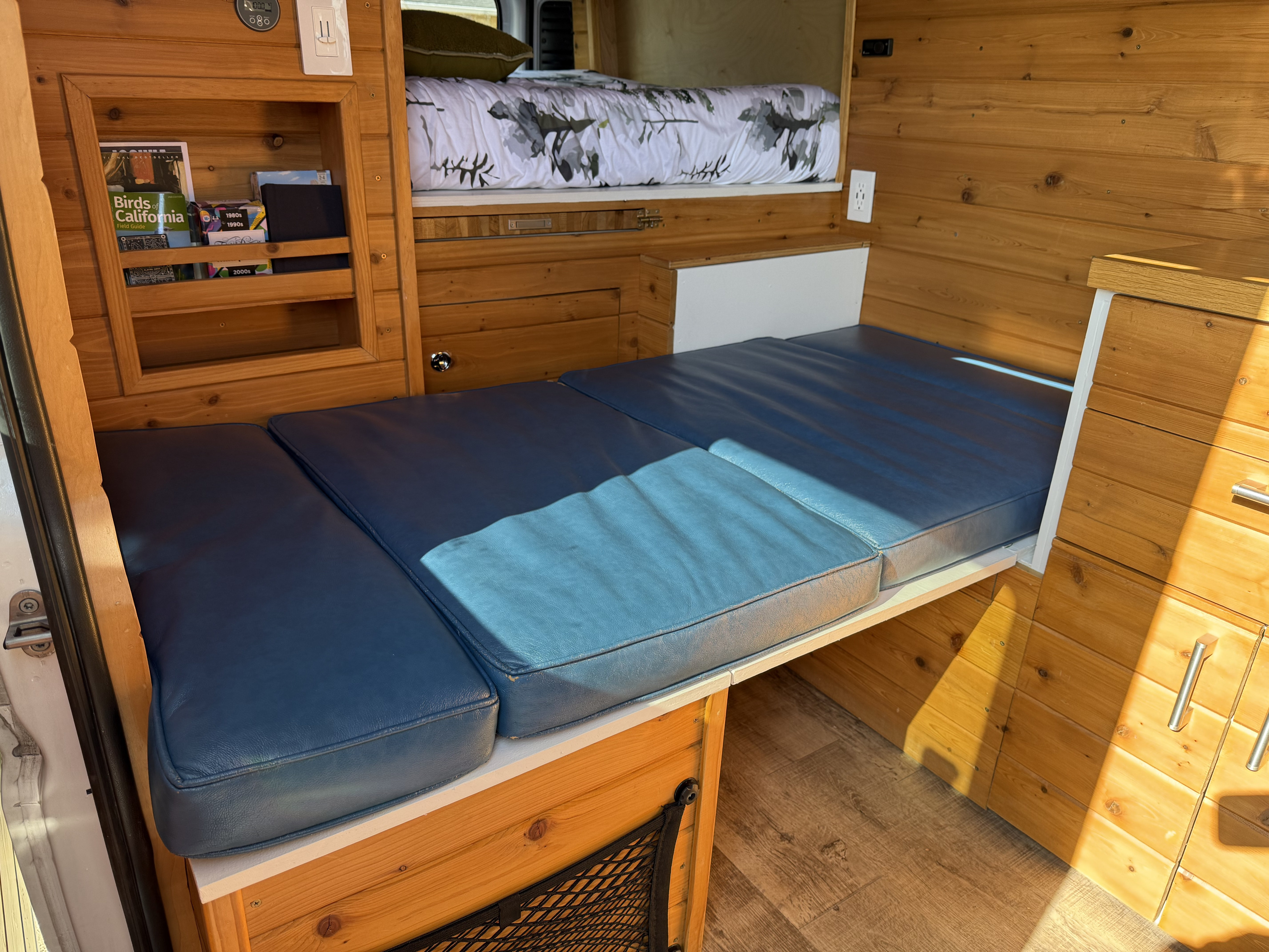 Cozy RV rental features wood walls, blue convertible seating, an overhead bunk bed, and storage for van life adventures.. RAM Promaster 2500 2020