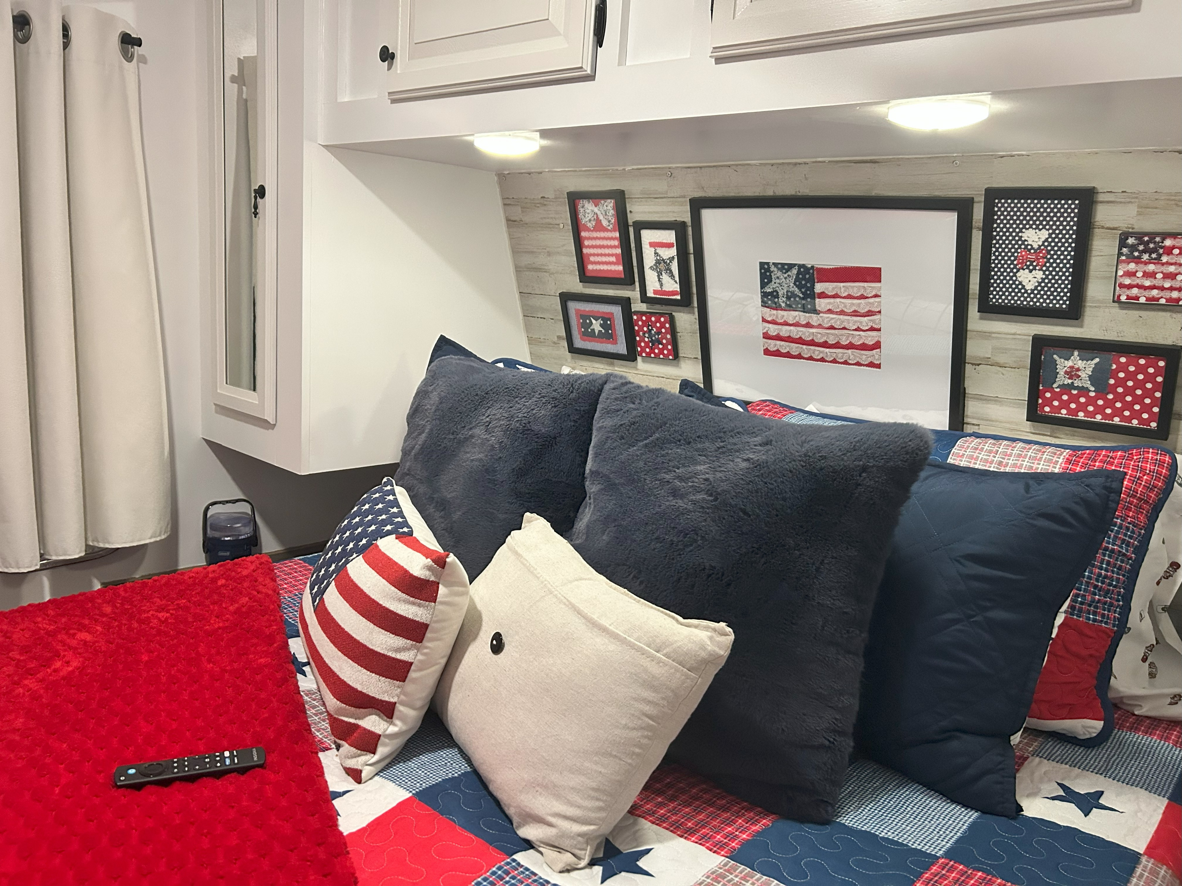 A cozy RV rental bedroom features a patriotic-themed bed with red, white, and blue pillows, framed flag art, and a TV remote on a red blanket.. Jayco Eagle 298RLDS 2013