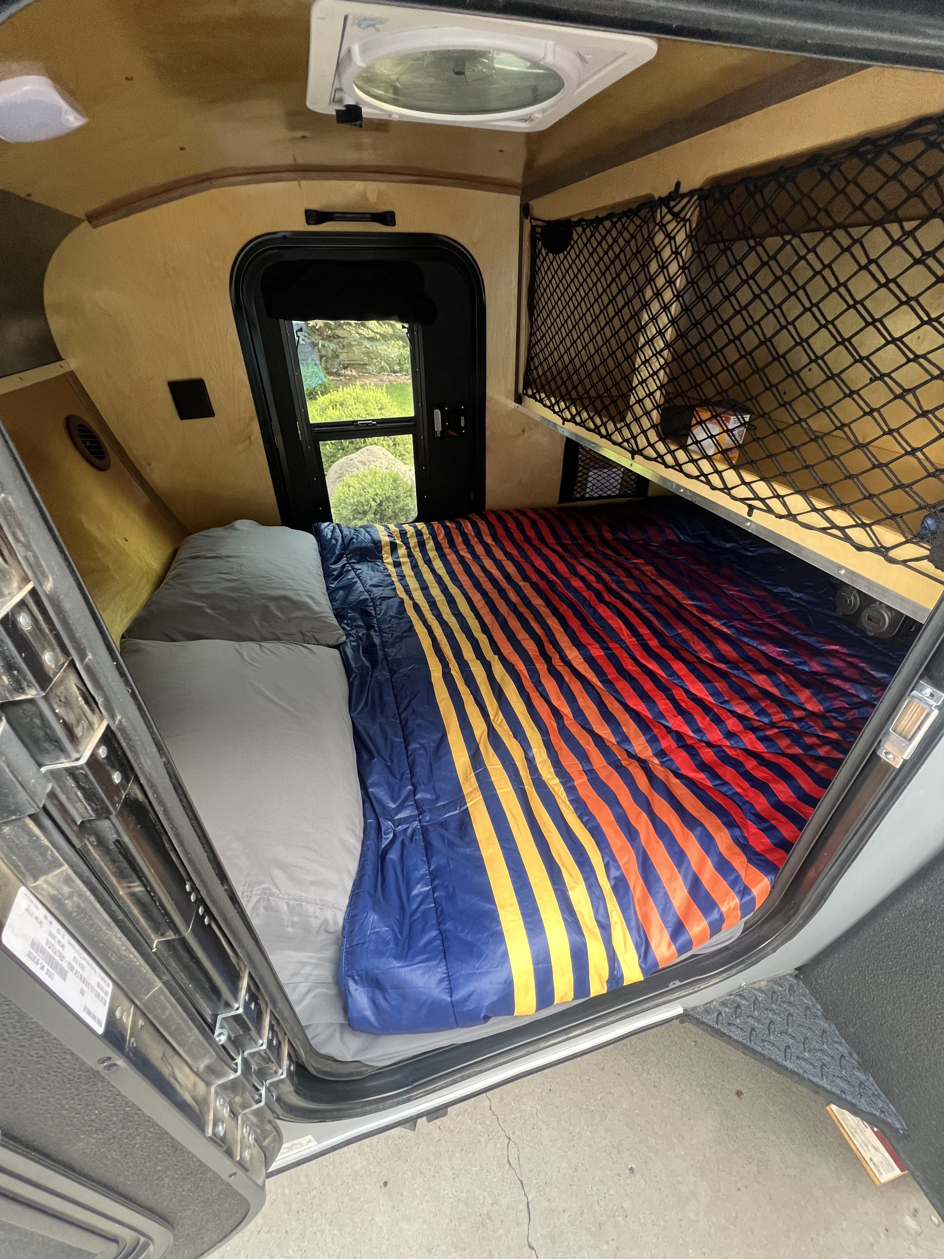 Cozy RV rental interior with a bed, colorful striped bedding, a window with outdoor view, and an overhead storage net.. Venture Trailer Works XTC 2025