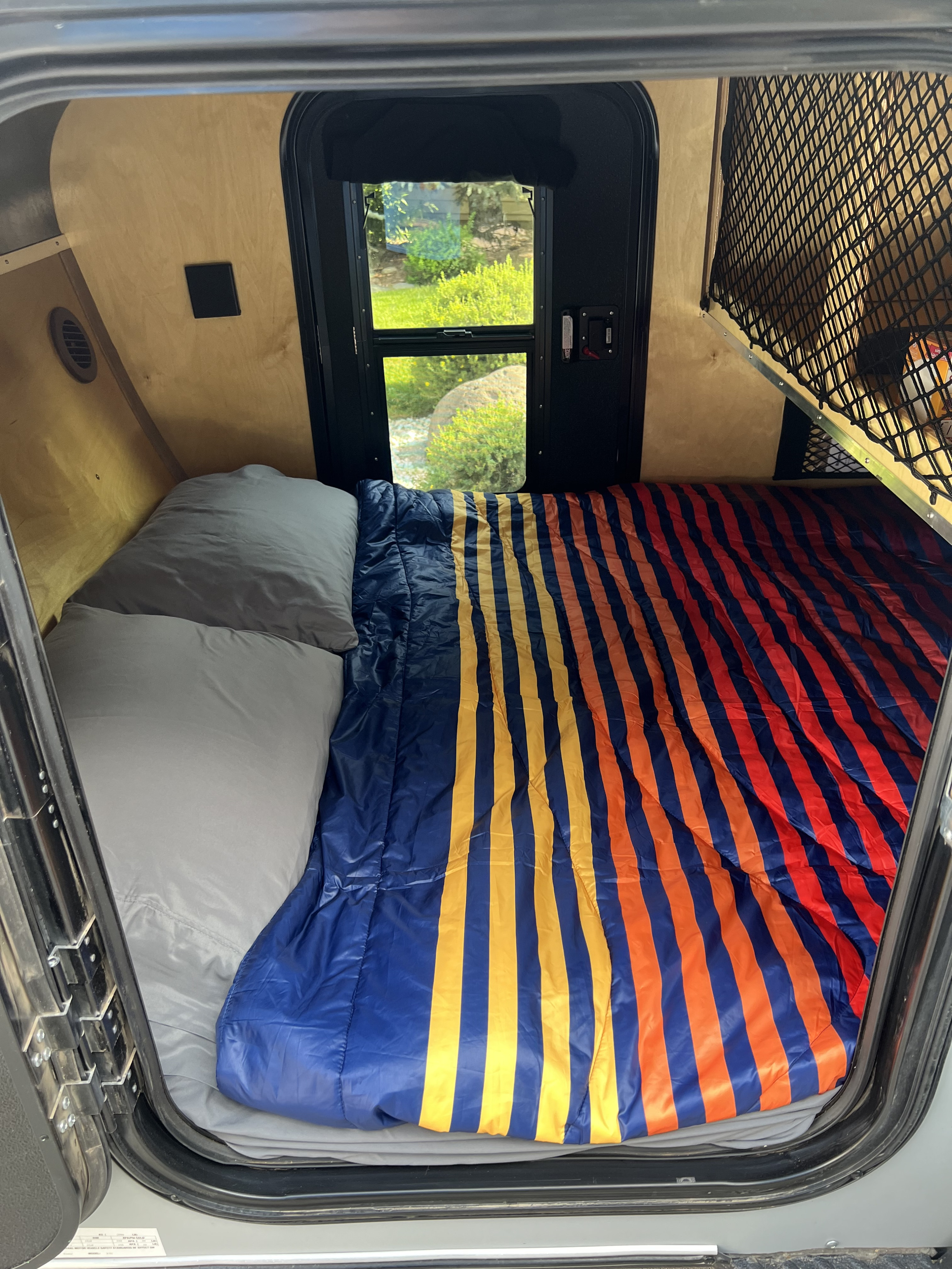 RV rental camper interior: cozy bed with vibrant striped comforter, pillows, and a refreshing window view of nature.. Venture Trailer Works XTC 2025