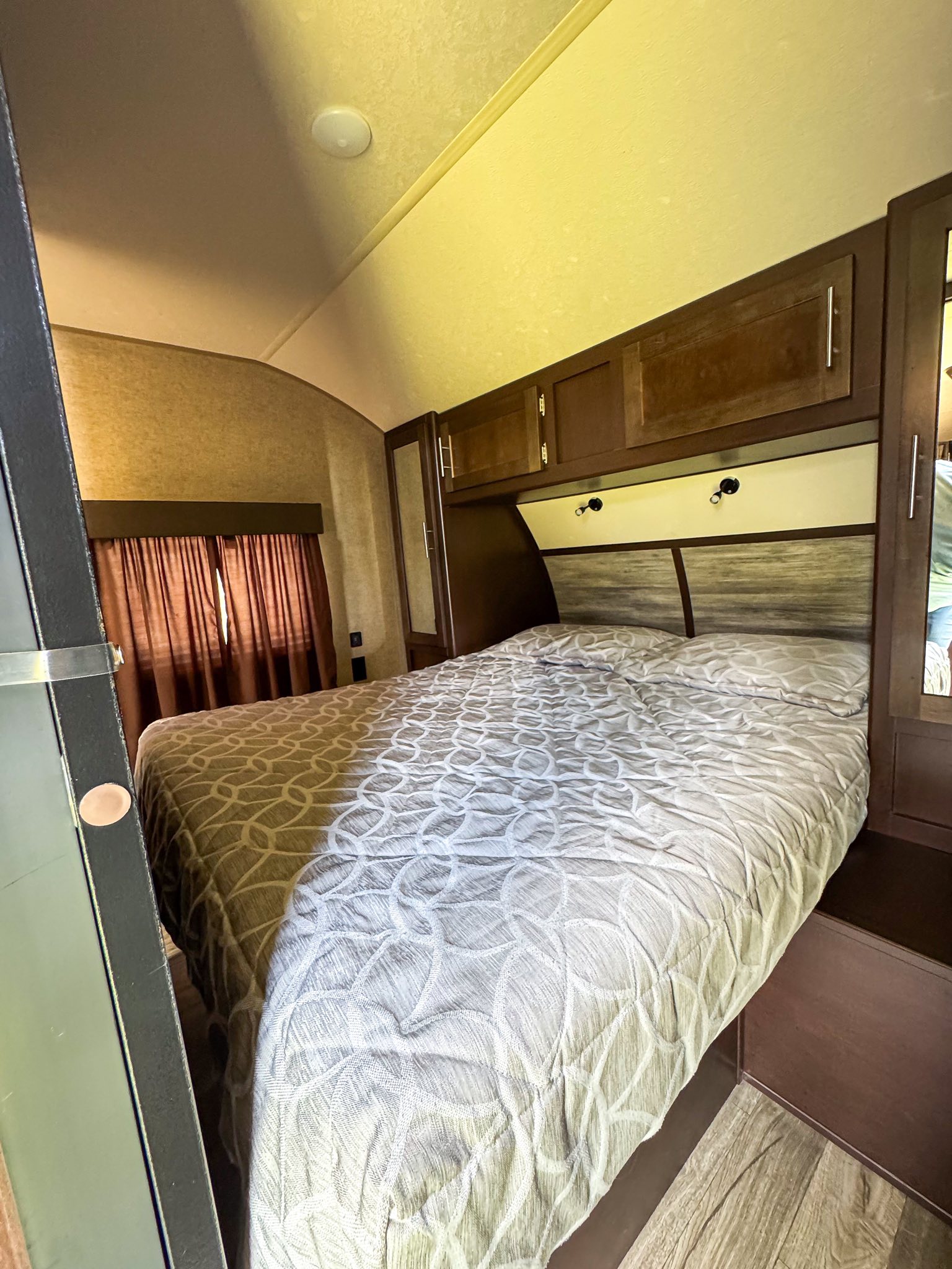 This RV rental bedroom offers a comfortable bed with patterned linens, dark wood cabinets, practical lighting, and a window with curtains.. Forest River Cherokee Grey Wolf 2017