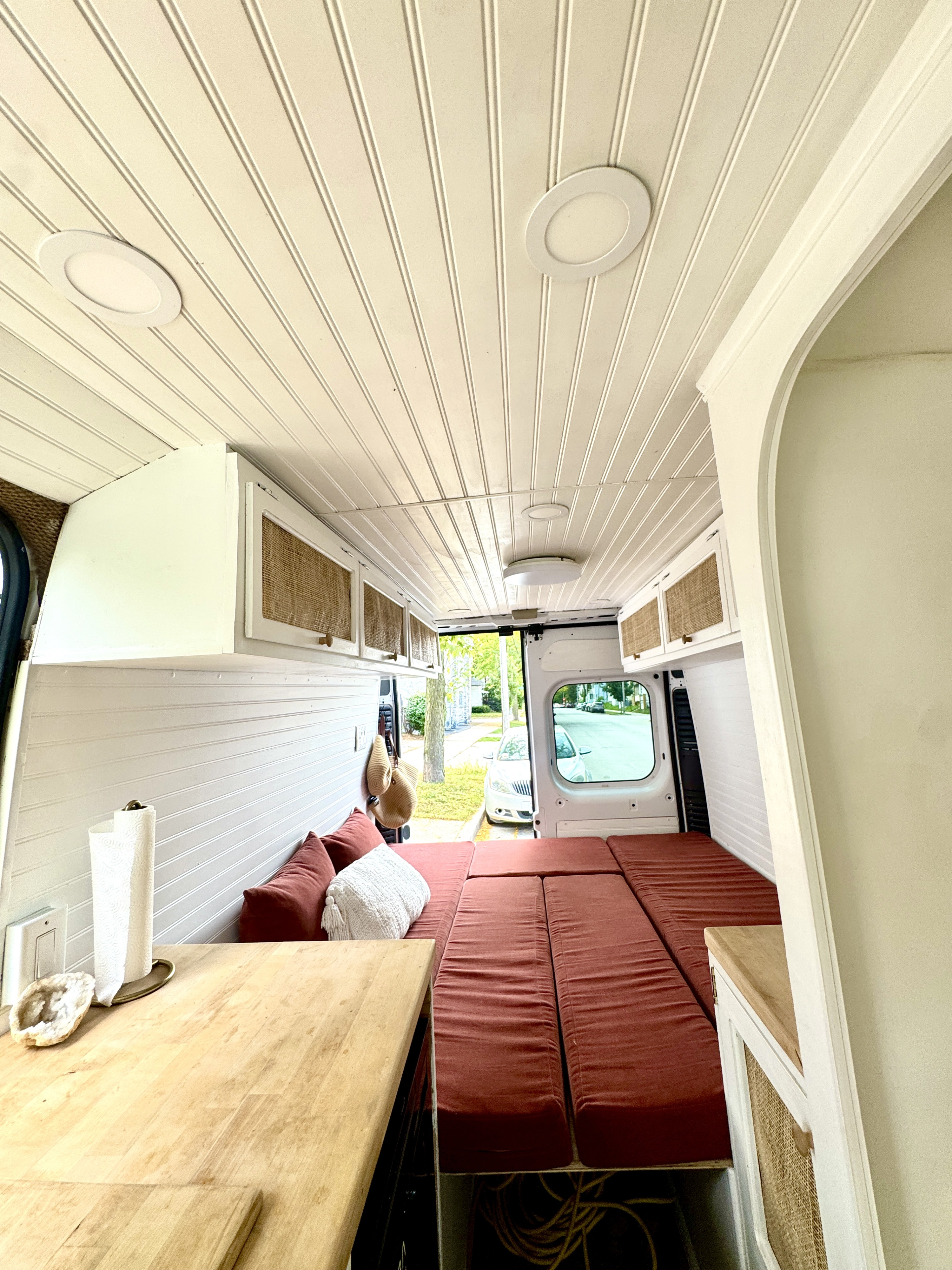 This RV rental's inviting interior features white beadboard walls/ceiling, rattan cabinets, and a comfortable red bed/lounge area.. Dodge Promaster 2019