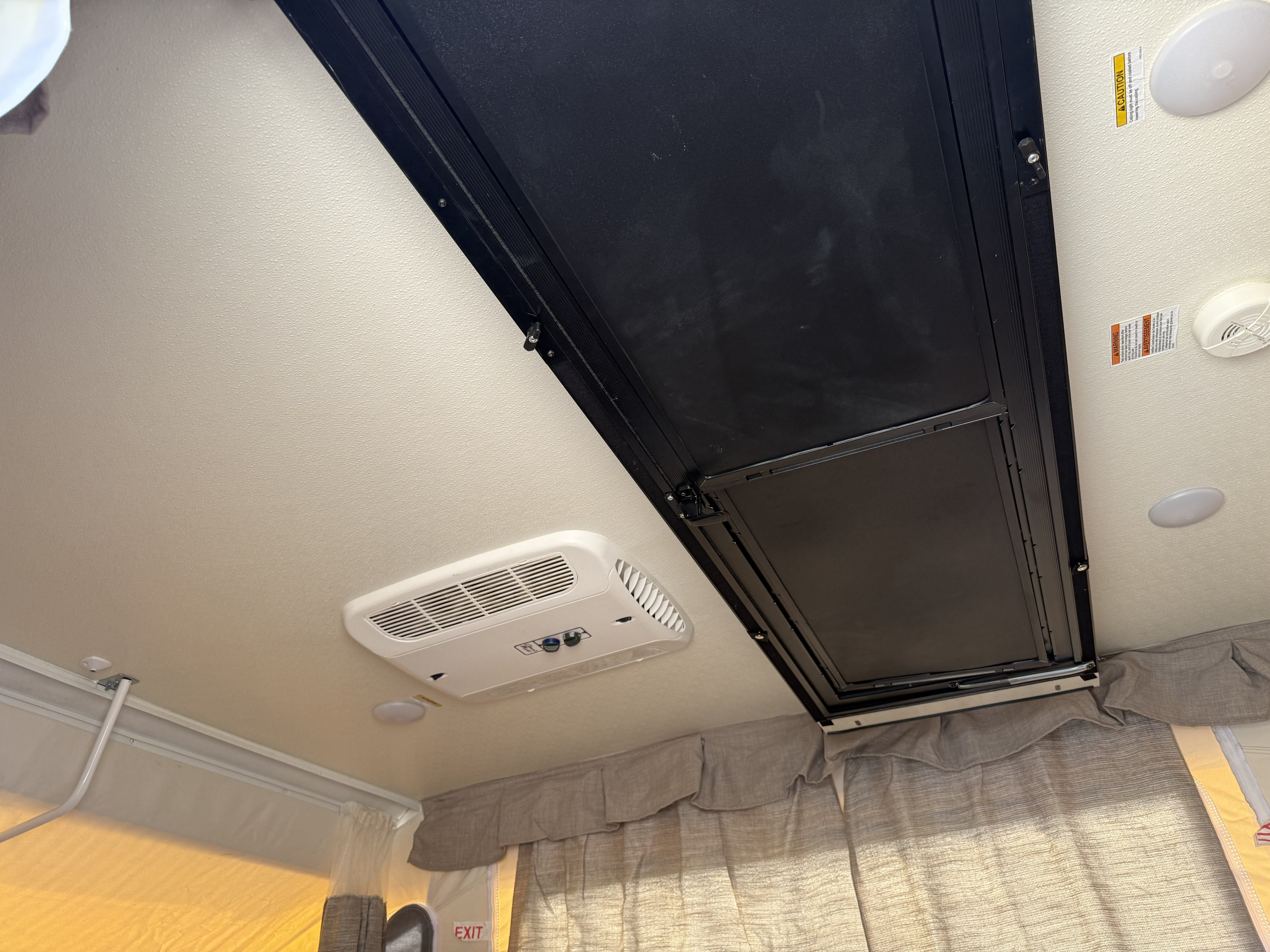 The interior ceiling of an RV rental pop-up camper, featuring an AC unit, ceiling lights, a black folding bed panel, and safety warning labels.. Forest River Flagstaff M.A.C. 2022