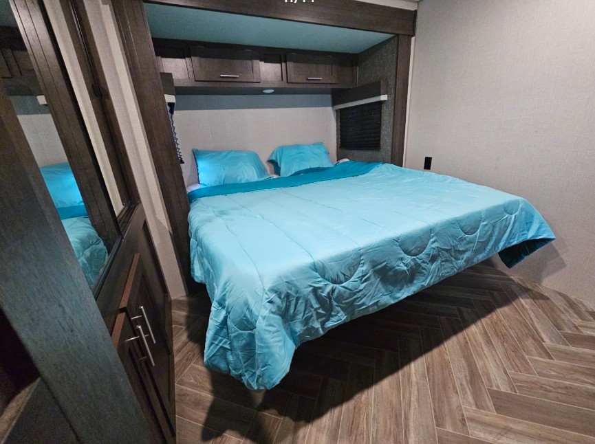 RV rental bedroom featuring a large bed with bright blue bedding, overhead cabinets, and wood-look herringbone floor.. KZ 362BH 2020