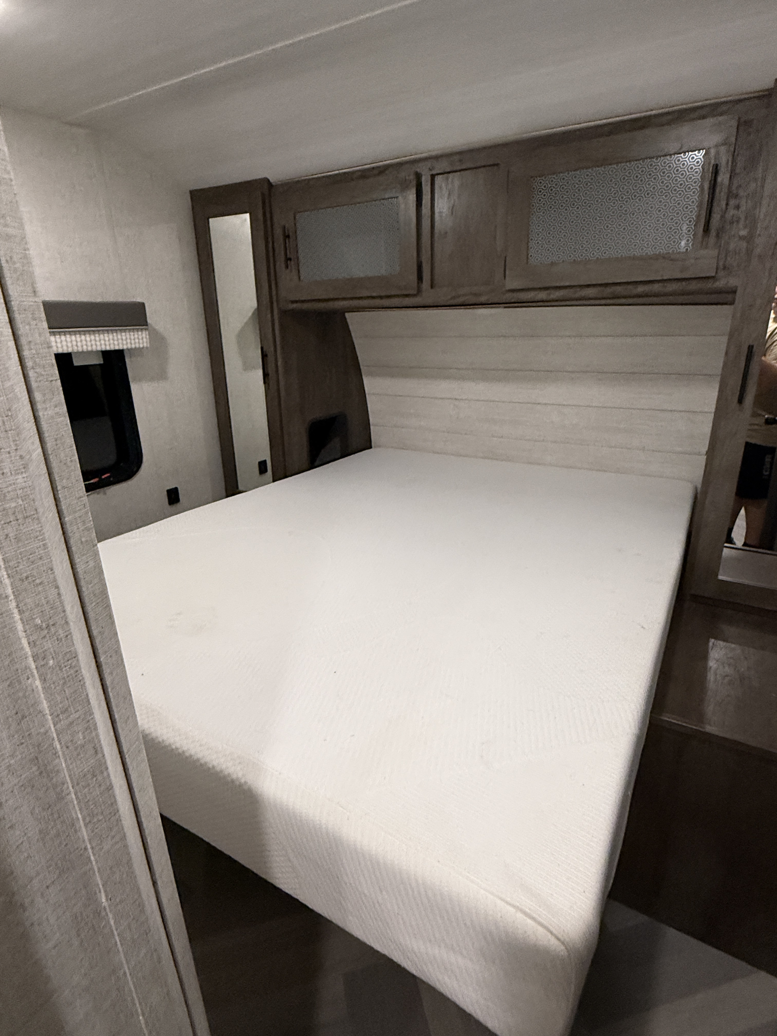 Clean RV rental bedroom featuring a white mattress, dark wood overhead cabinets, a mirrored wardrobe, and a small window.. Forest River Salem 2021