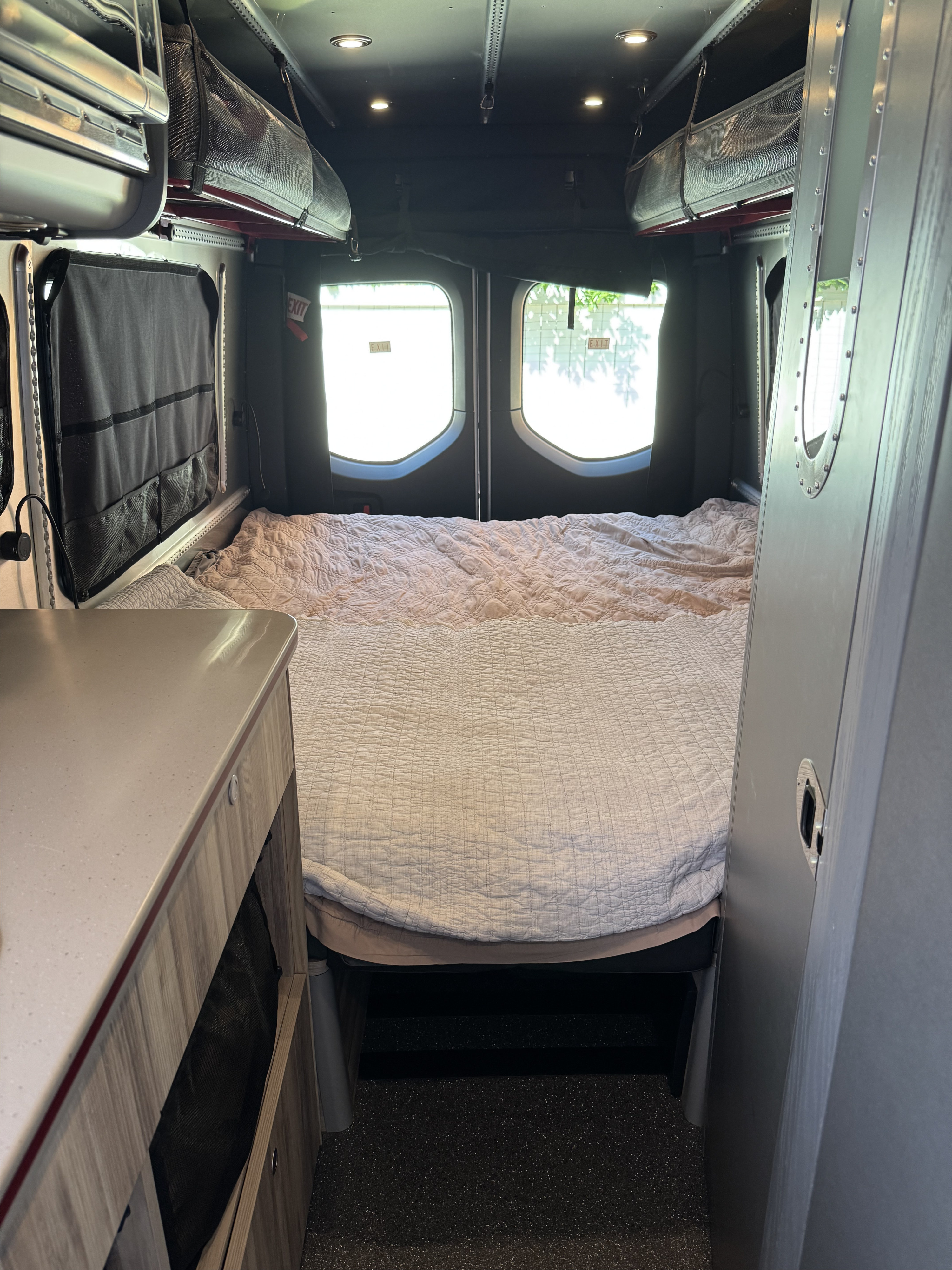 Cozy RV rental camper van bedroom. Features a made bed, overhead storage, and view out the rear doors.. Airstream Interstate 2023