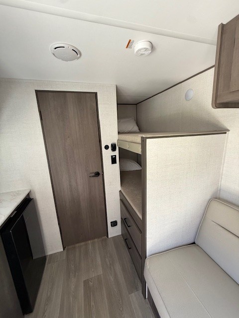 RV rental interior: visible top bunk bed, light sofa, wooden door, and floor. Ceiling has a vent & smoke detector.. Grand Design Transcend One 2025