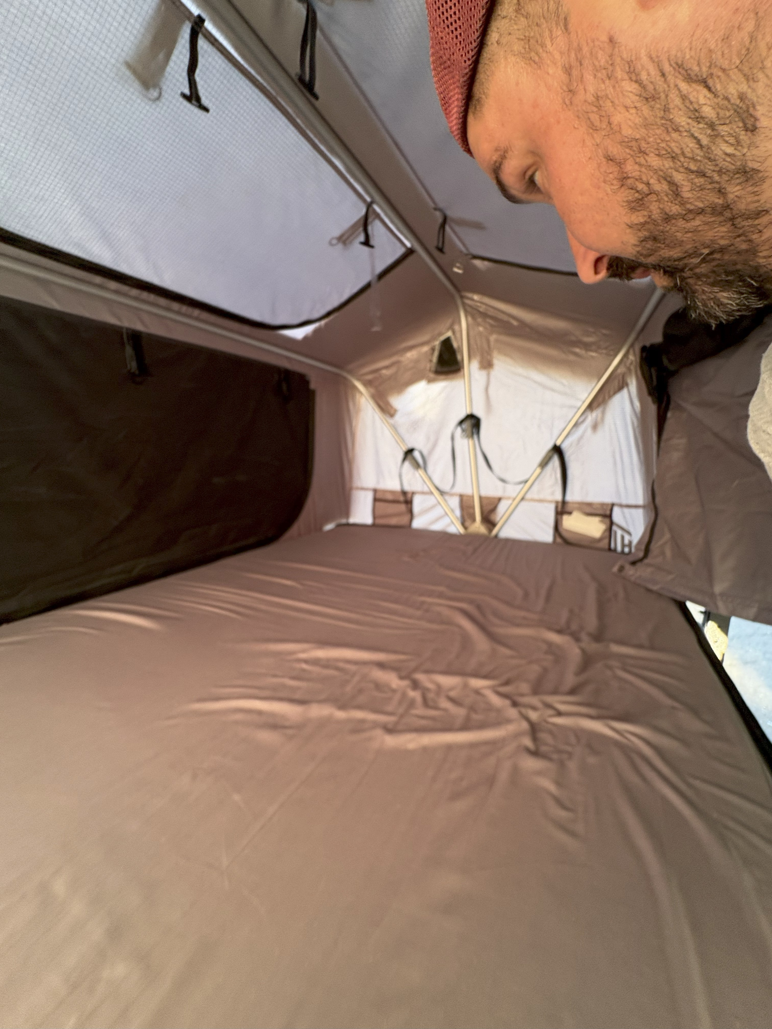 Inside an RV rental's pop-up tent, showing a comfortable sleeping area with a neatly made bed and storage pockets. A man peers in from above.. Silver Eagle Manufacturing M1102 2013