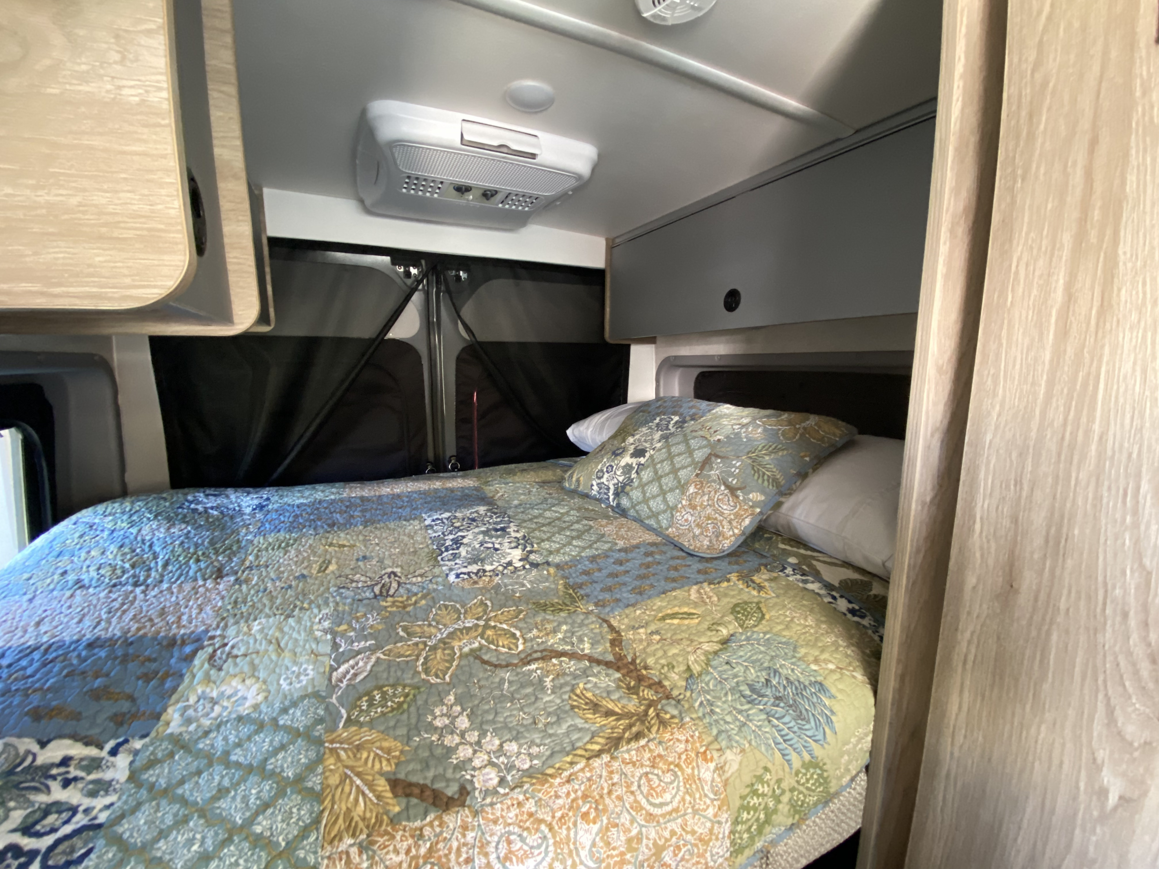 Cozy RV rental bedroom with a colorful quilt, overhead cabinets, and AC unit for comfortable travels.. Winnebago Solis Ram 2025