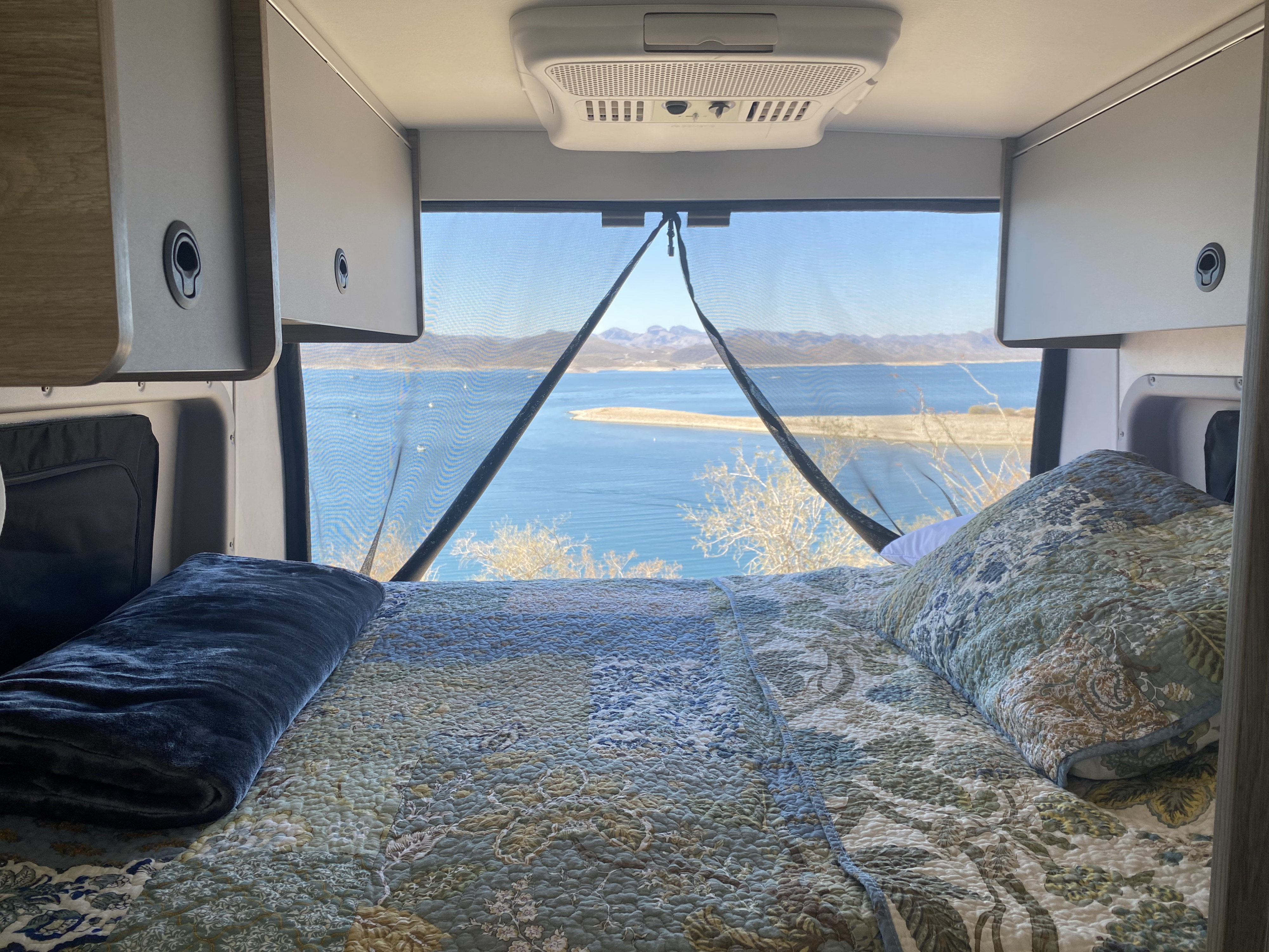 Cozy **rv rental** interior: a comfortable bed, overhead cabinets, and AC, with a stunning lake and mountain view through a mesh screen.. Winnebago Solis Ram 2025