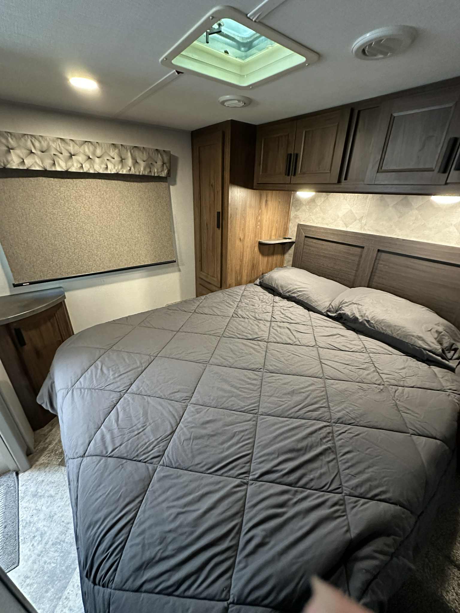 Comfortable RV rental bedroom with gray quilted bedding, wooden headboard, overhead cabinets, and a window blind.. Forest River Sunseeker LE 2021