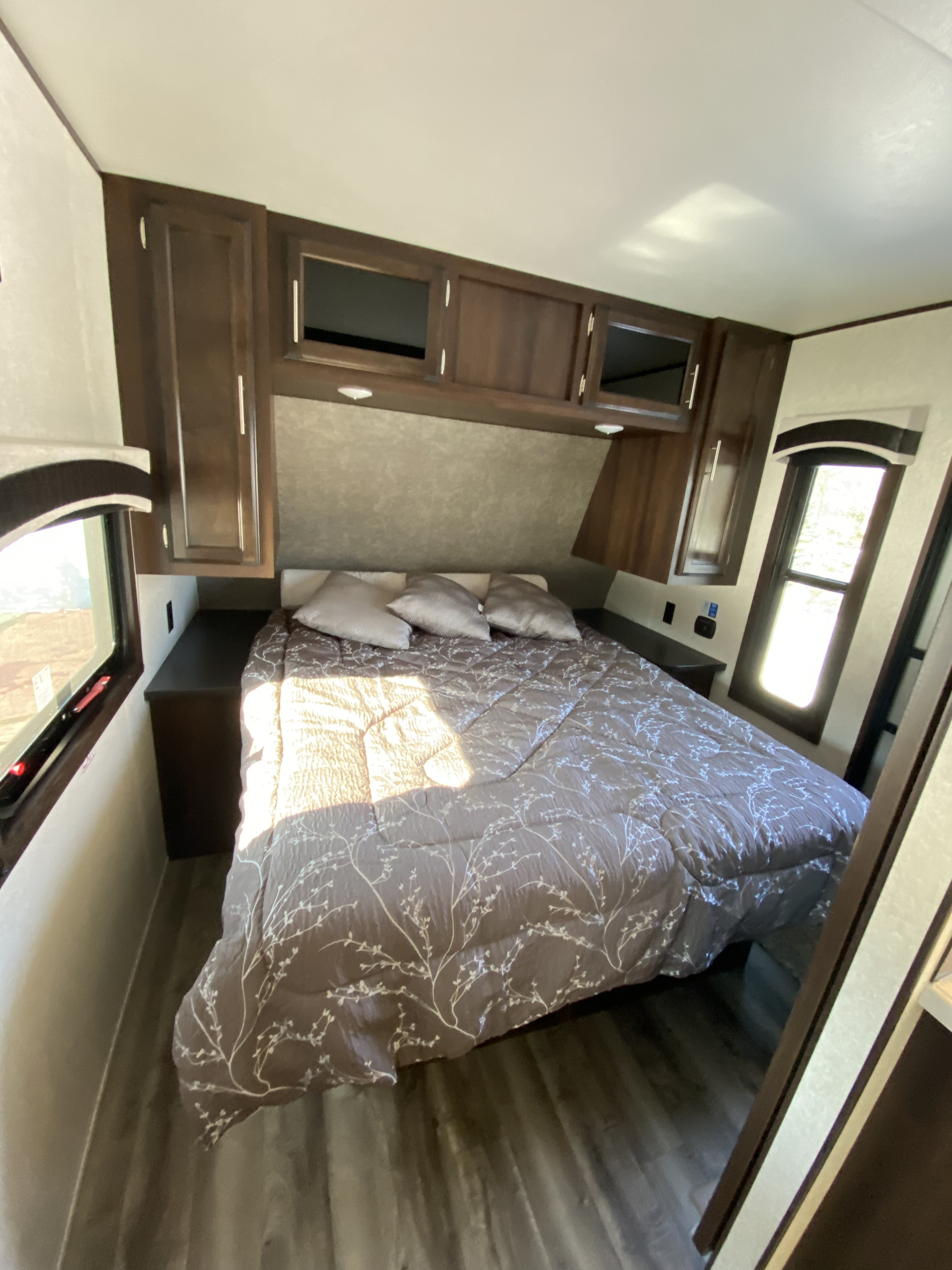 Cozy RV rental bedroom: a comfortable bed with patterned comforter, dark wood overhead cabinets, and sunlit side windows.. Jayco Jay Flight 2019