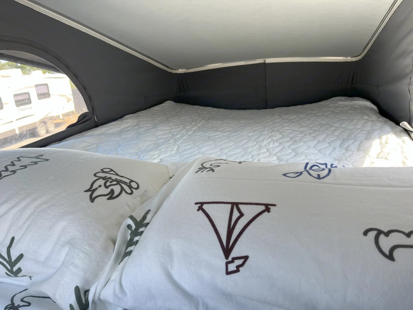 Inside a cozy pop-top rv rental: comfortable bed with unique patterned pillows. Another RV is visible outside the window.. Winnebago Solis 2023