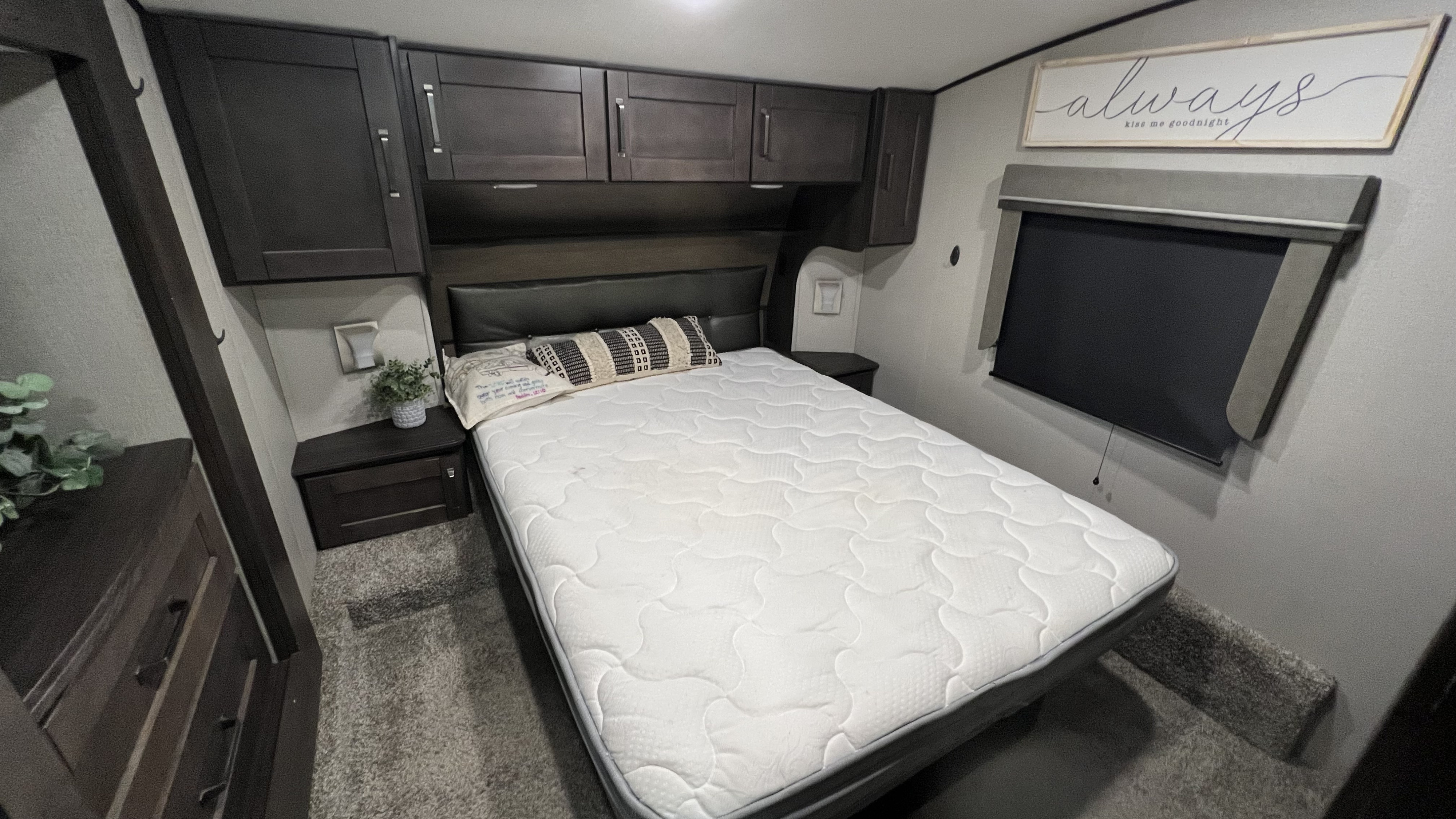 Cozy RV rental bedroom featuring a white quilted mattress, dark overhead storage, nightstands, and a window blind.. Grand Design Reflection 2020