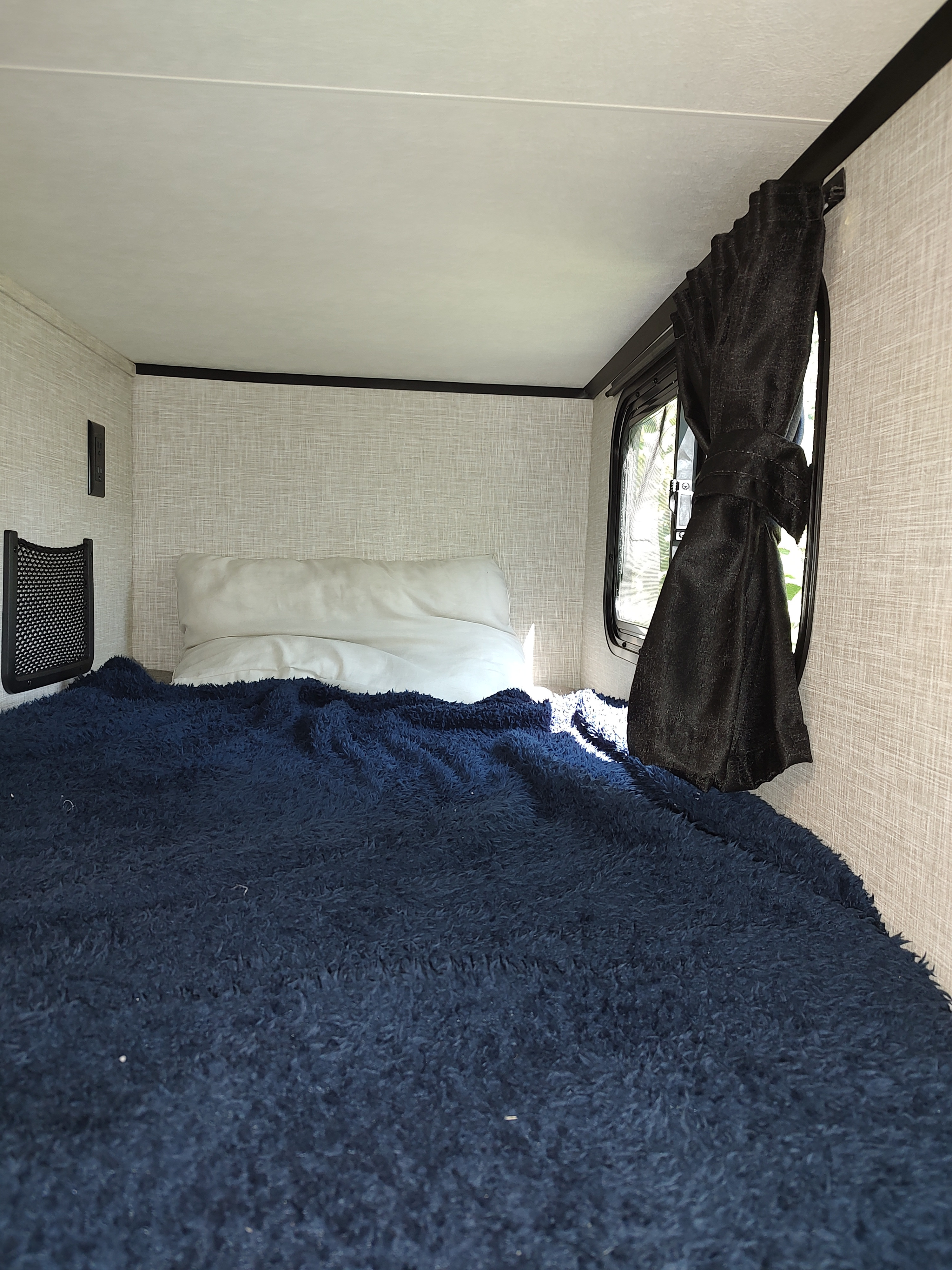 Inside an RV rental: a snug sleeping nook with a dark blue fluffy blanket, white pillow, and window with a tied black curtain.. Jayco Jay Feather Air 18mbh 2024