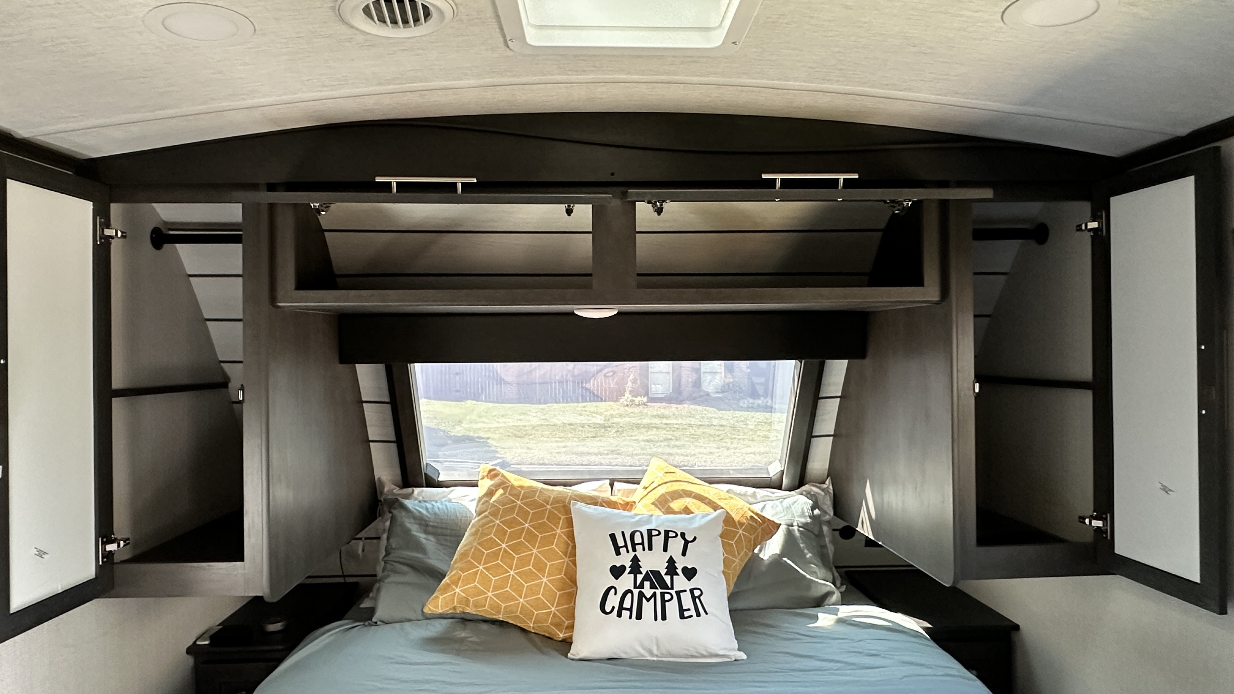 RV rental bedroom interior with a cozy bed, "Happy Camper" pillow, overhead storage, and side wardrobes. Bright window view.. Keystone Cougar Half Ton (22MLS) 2025