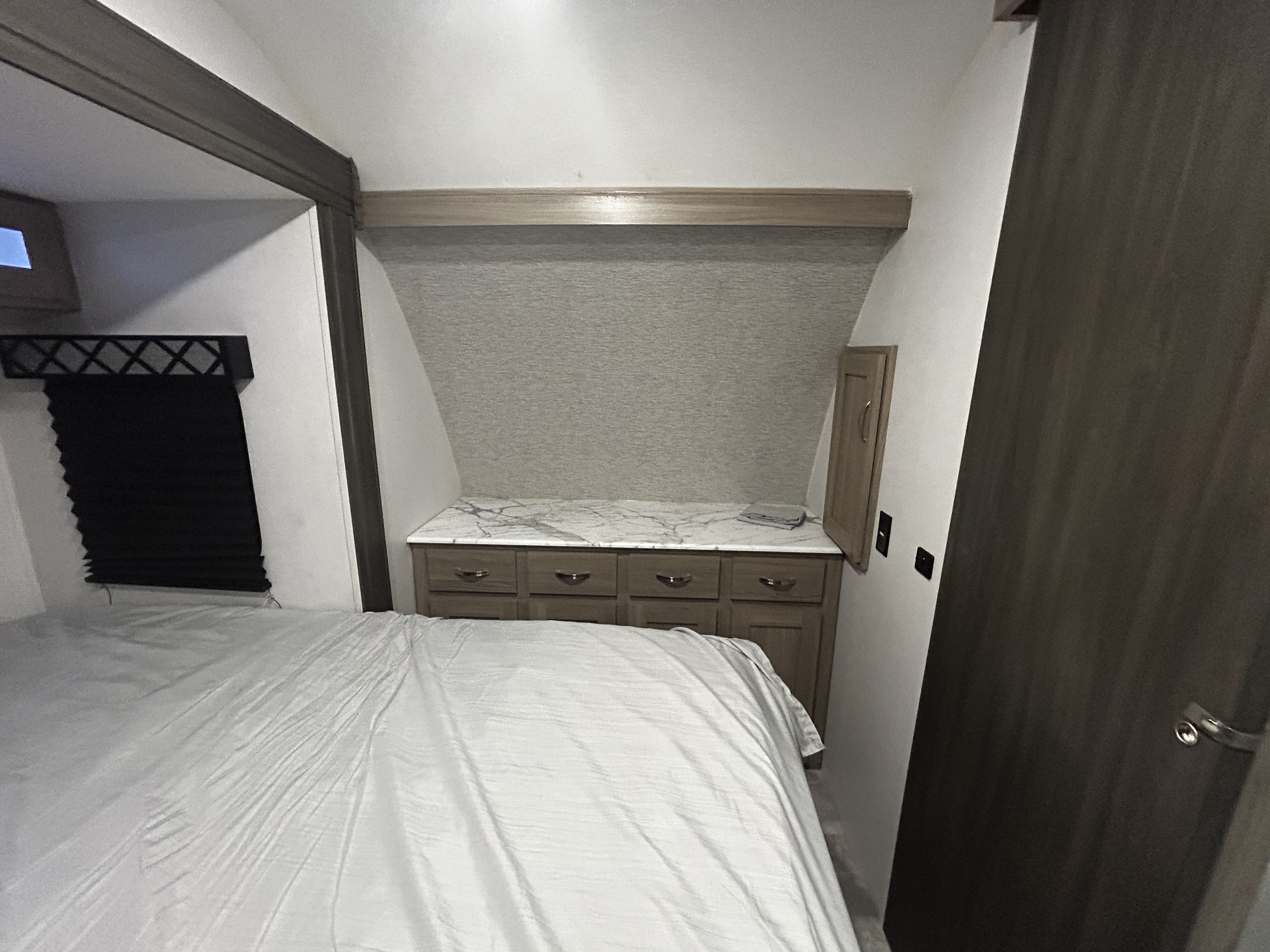 RV rental bedroom with a comfy bed, built-in storage dresser, marble-look counter, and a dark pleated window blind.. Work and Play Toy Hauler 2022