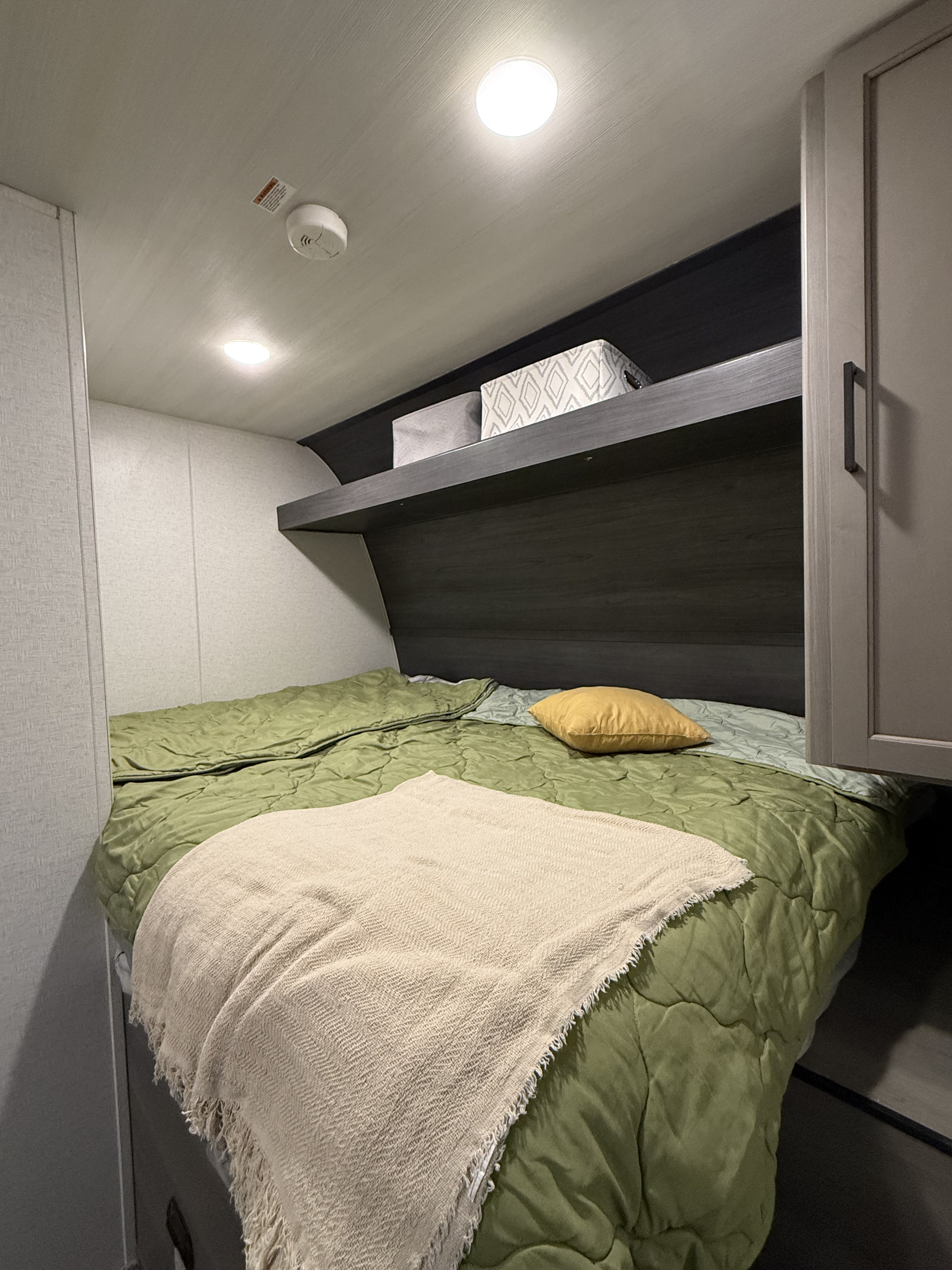 This RV rental bedroom offers a comfortable bed with green and beige bedding, a yellow pillow, and overhead storage.. Keystone RV Hideout Sport 2024