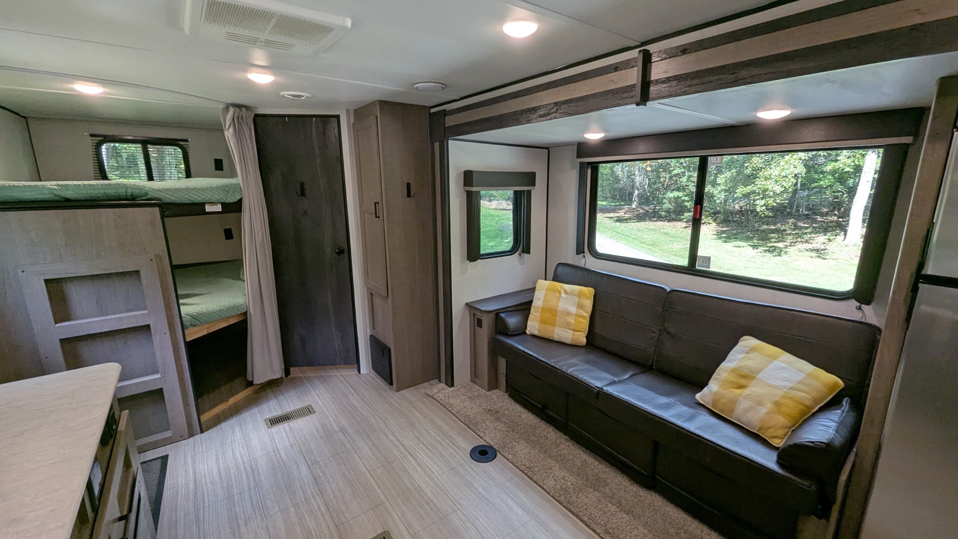 Bright rv rental interior featuring bunk beds, a dark couch with yellow pillows, and large windows offering a tree view.. Keystone Hideout 2021