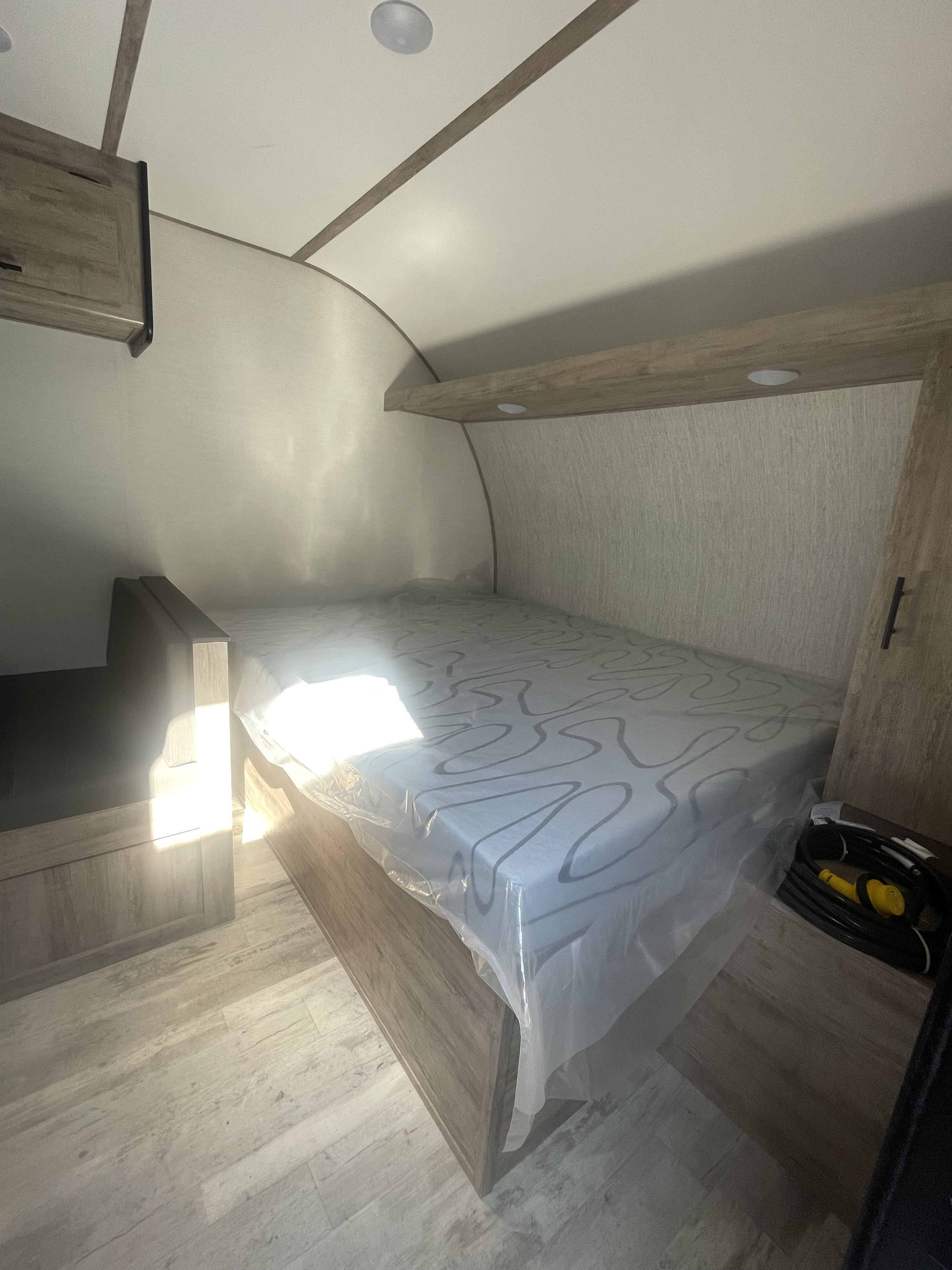 RV rental bedroom with a new, plastic-covered mattress, wooden accents, overhead shelf with lights, and light flooring. Sunlit and clean.. Gulf Stream Grand River 2026