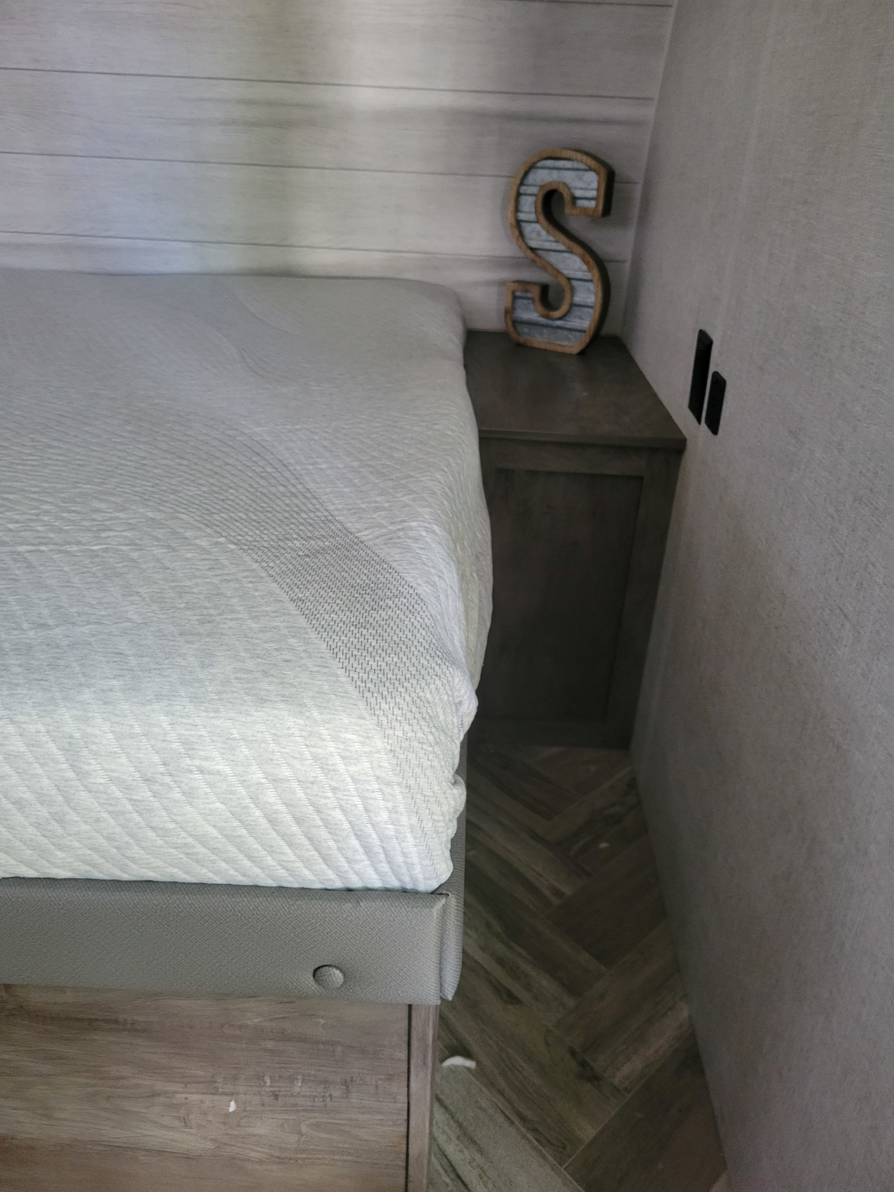 Cozy bed and nightstand in an RV rental, featuring wood paneling, decorative 'S', and herringbone floor.. Forest River Salem FSX 2021