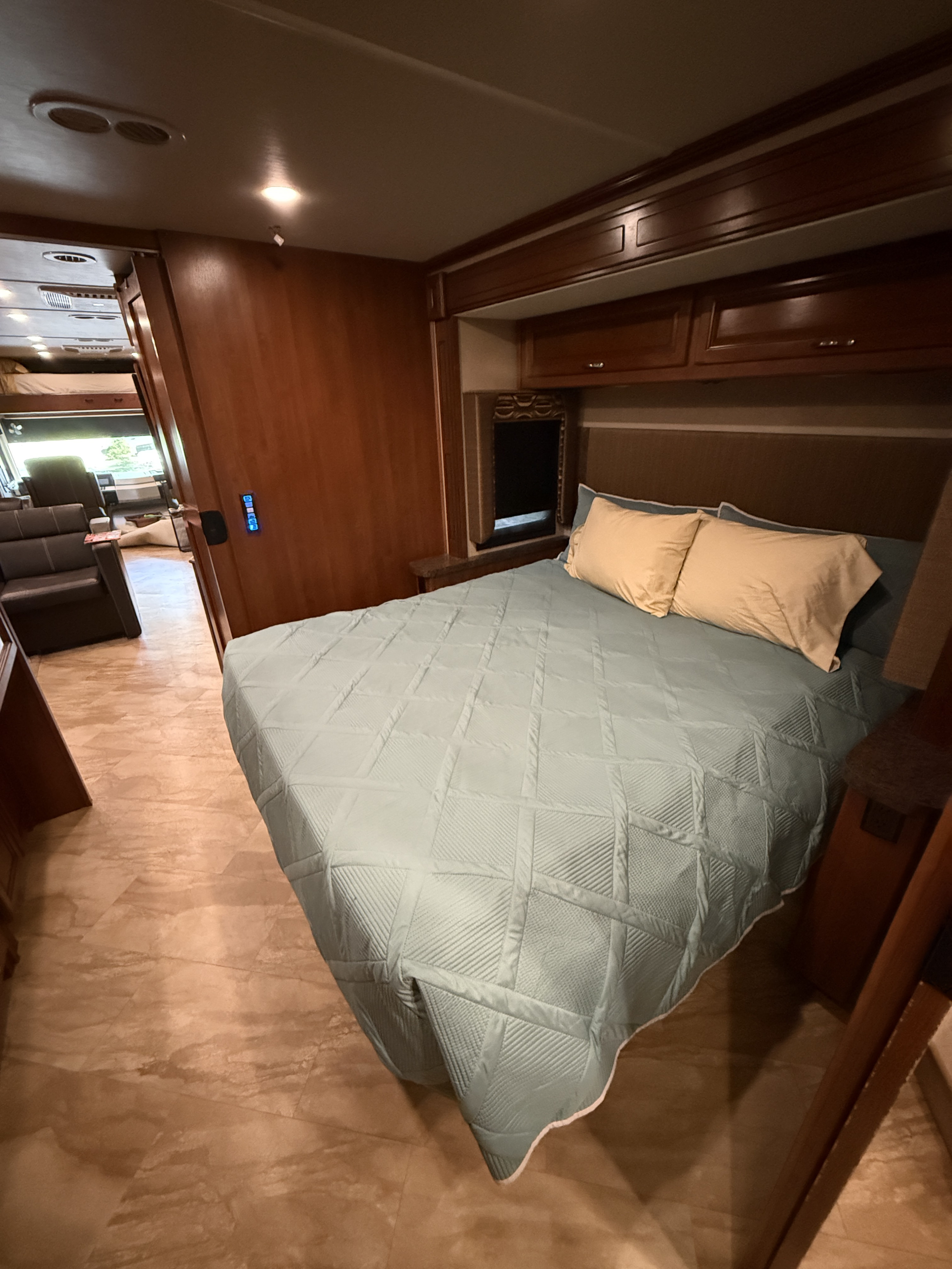 This inviting RV rental bedroom features a cozy bed with a light blue quilt, wooden cabinetry, and a glimpse into the living area.. Fleetwood Bounder 2016