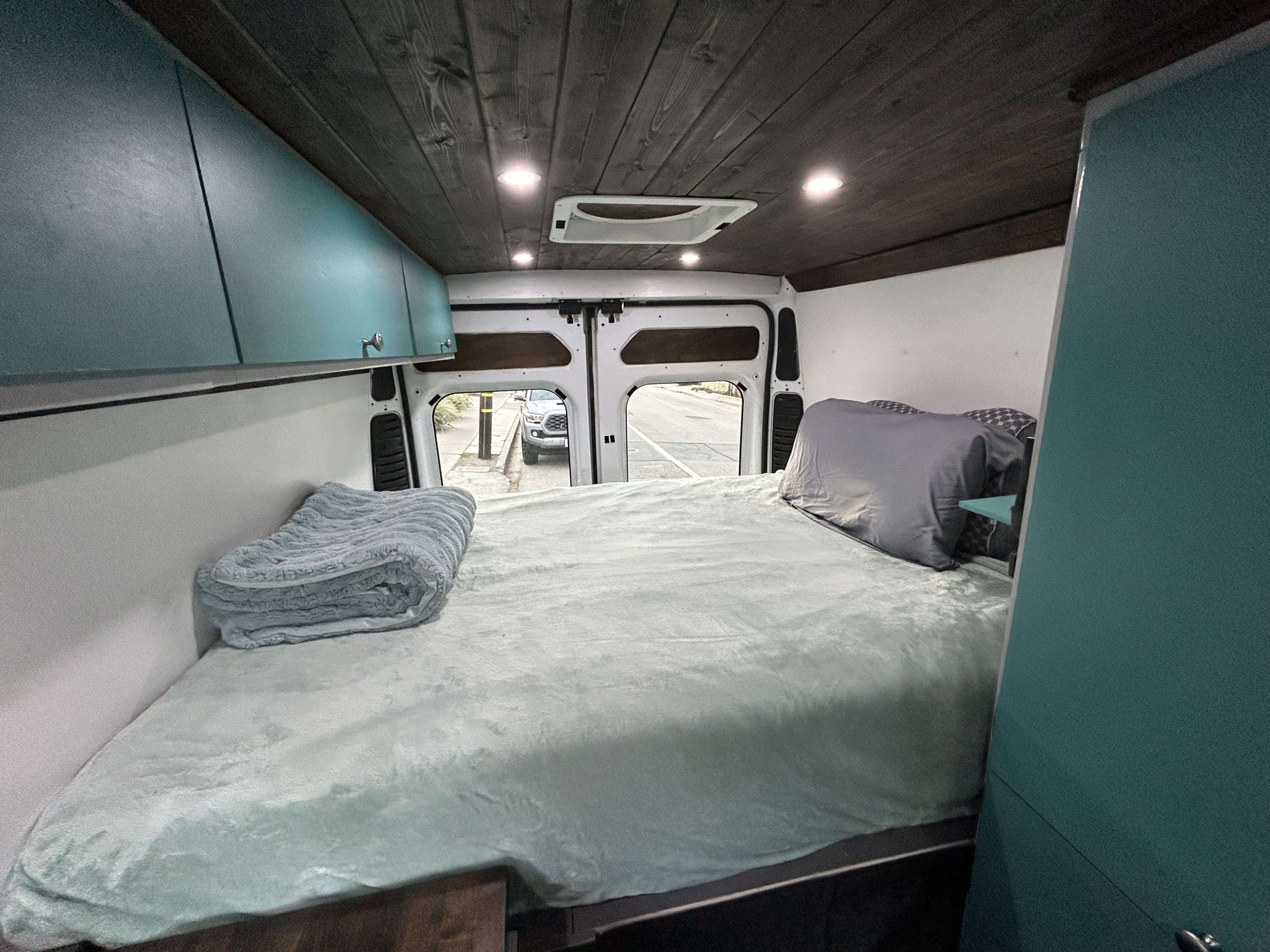 Inside a cozy rv rental camper van: large bed with teal bedding, overhead cabinets, wood plank ceiling, and open rear doors.. Ram Promaster 3500 2018