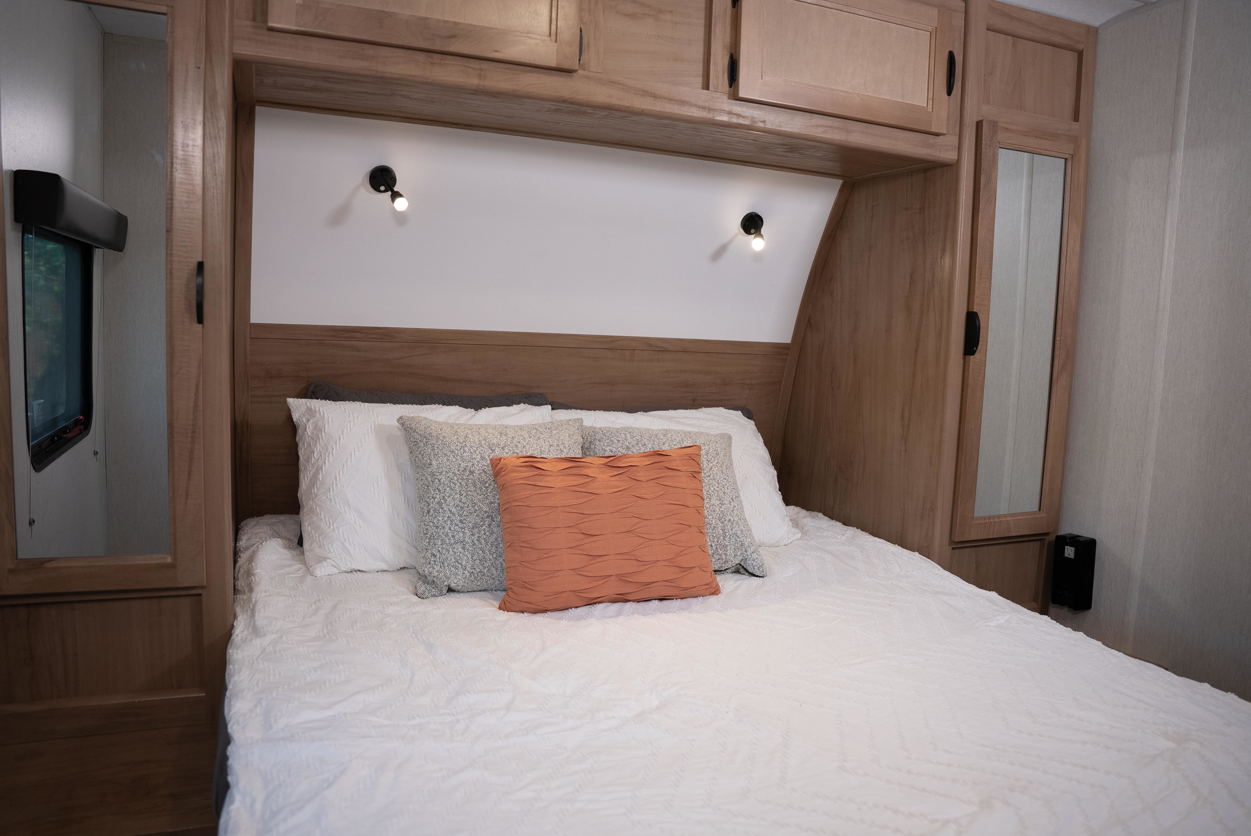 Interior view of an RV rental showing a bed with white bedding, accent pillows, wood cabinets, and reading lamps.. Forest River Black Label 2024
