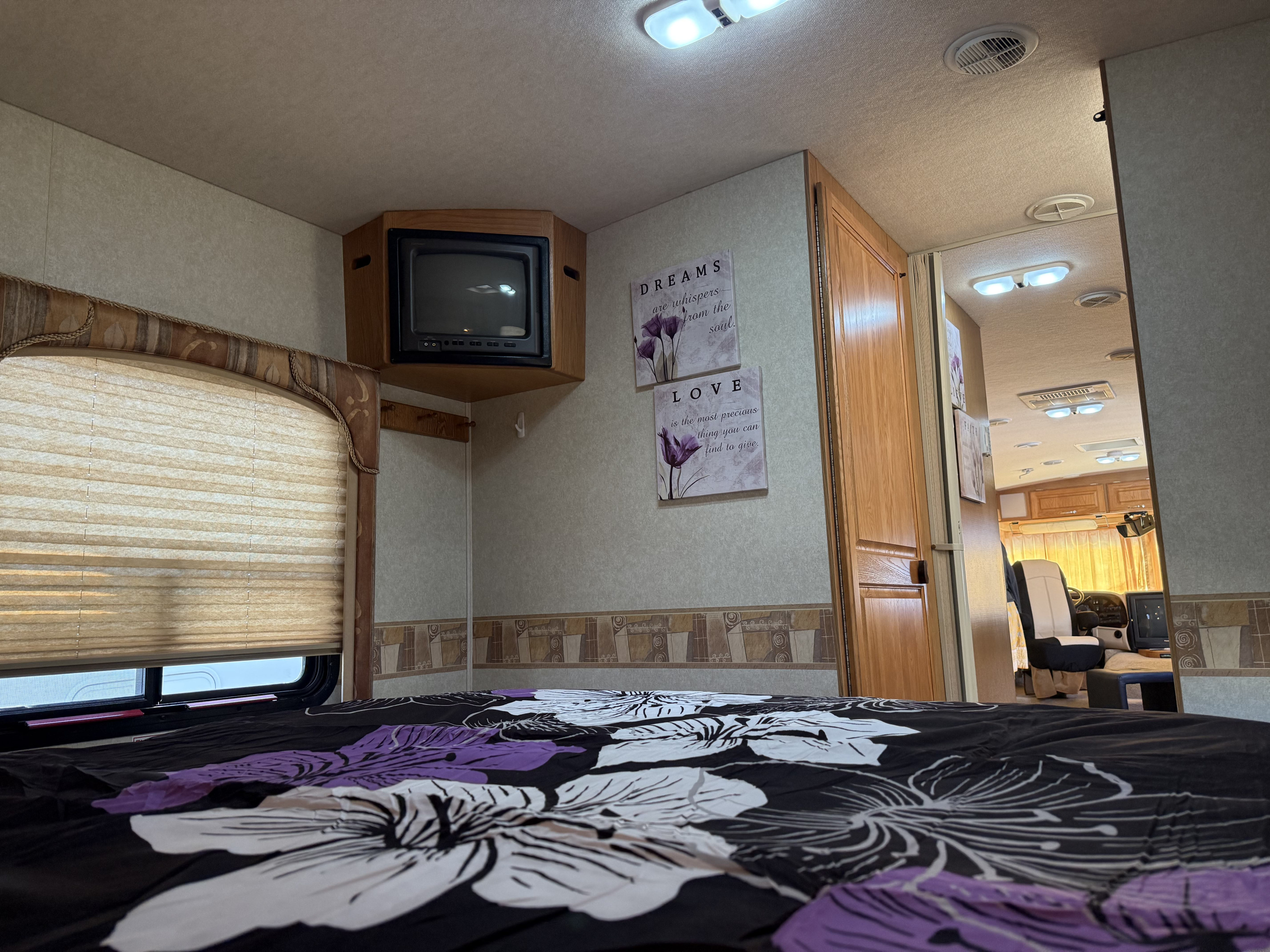 Cozy RV rental bedroom with floral bedding, wall art, a small TV, and an open view to the living area beyond.. Triple E Embassy 2003
